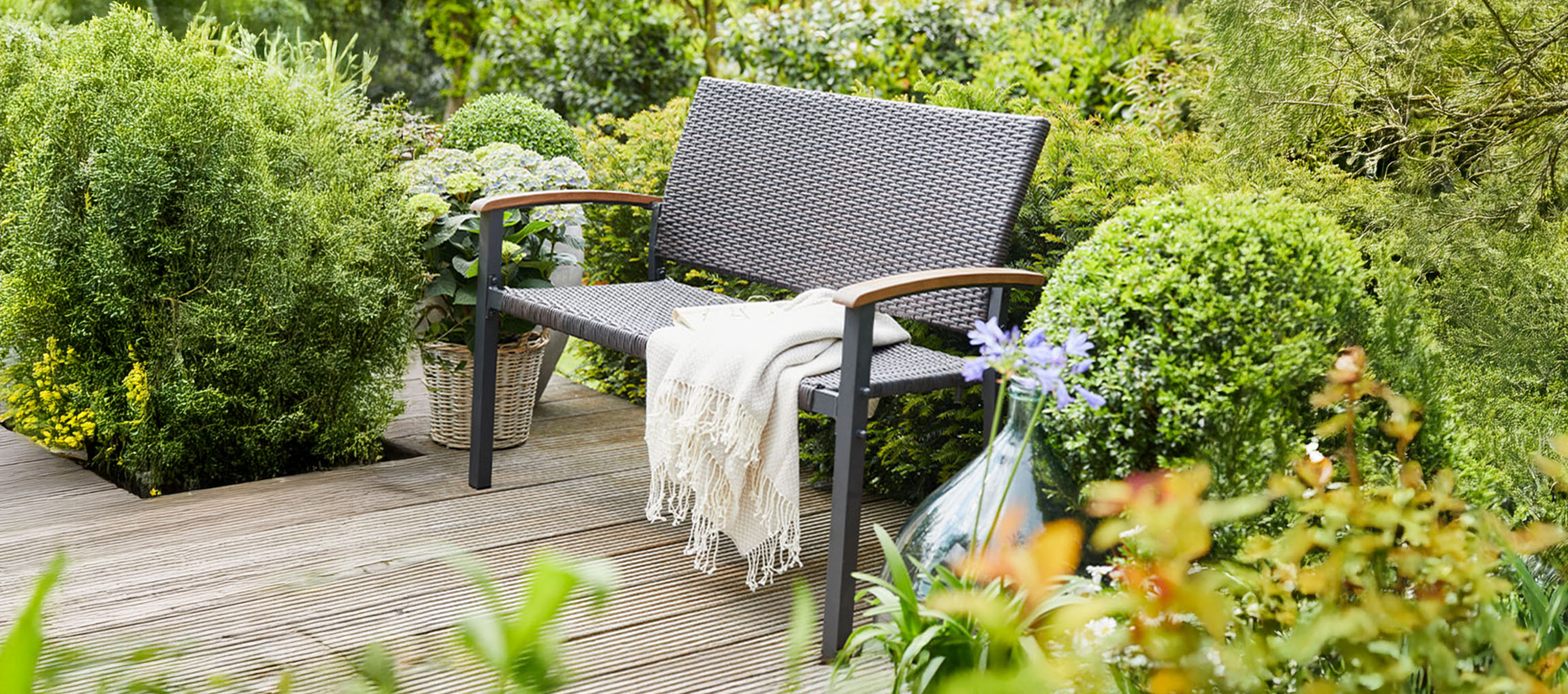 Un banc de jardin gris avec une couverture sur une terrasse en bois entourée de plantes vertes.