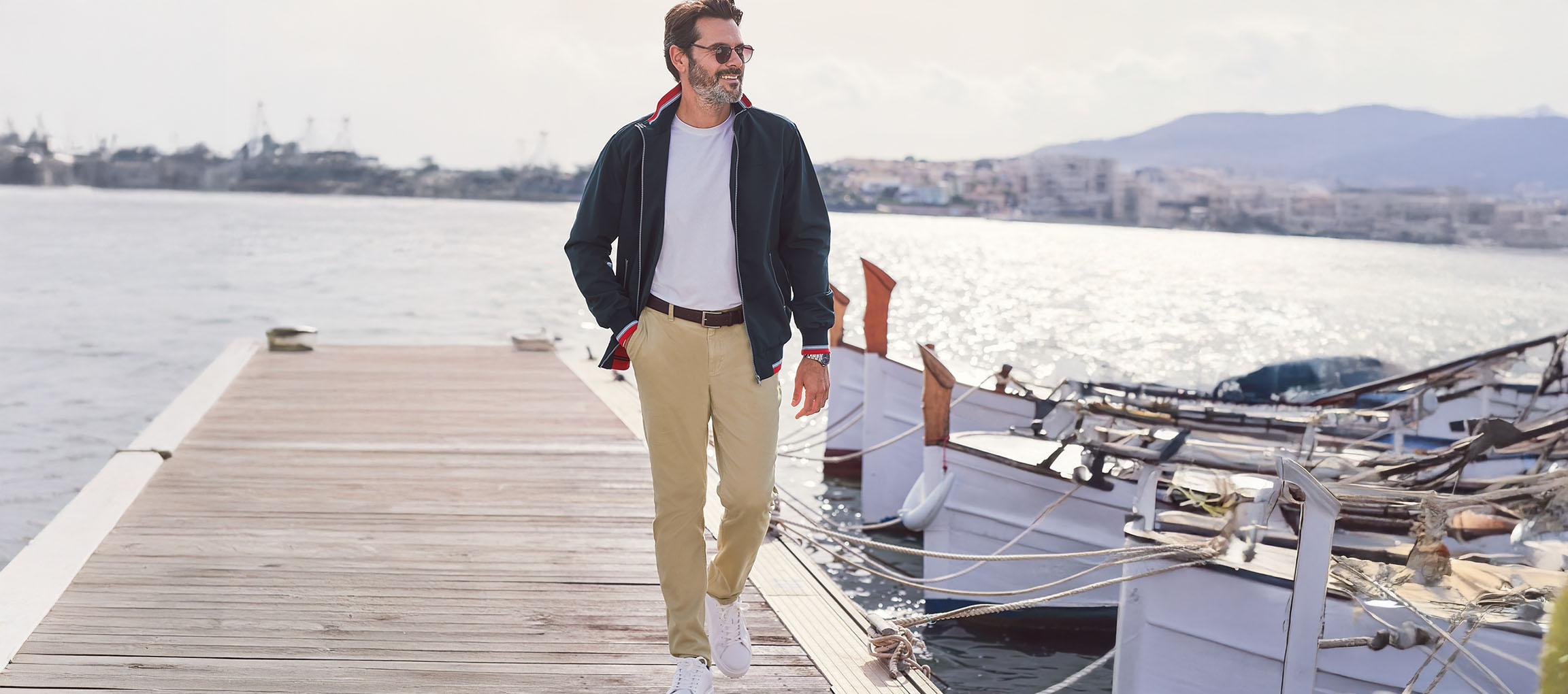 Homme en blouson aviateur vert foncé, pantalon chino et baskets blanches sur un quai.