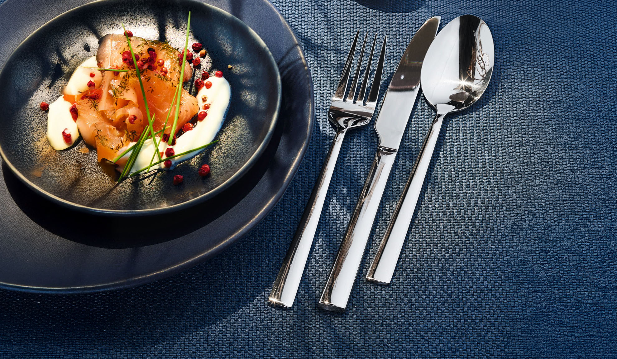 Saumon fumé avec crème, herbes et baies roses sur une assiette foncée avec couverts.