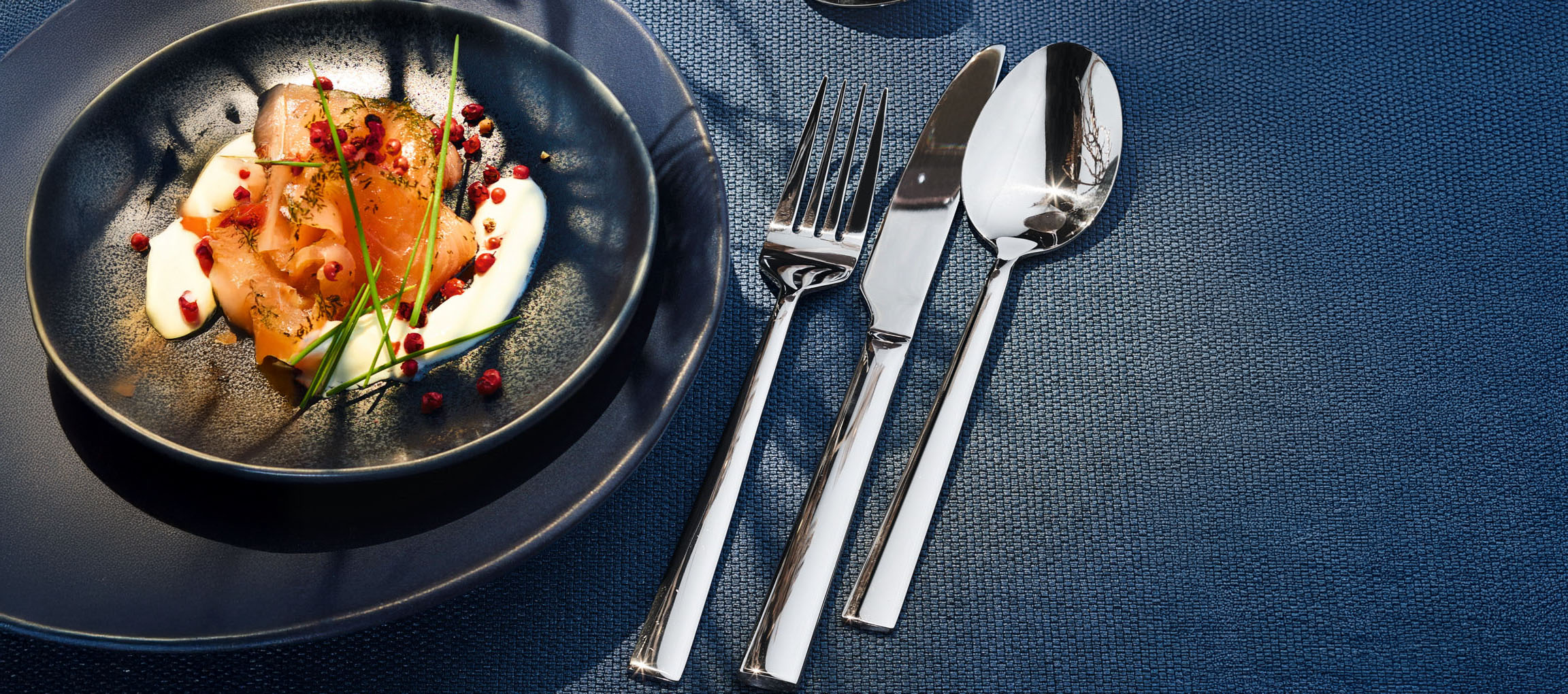 Saumon fumé avec crème, ciboulette et poivre rose sur une assiette foncée avec couverts.