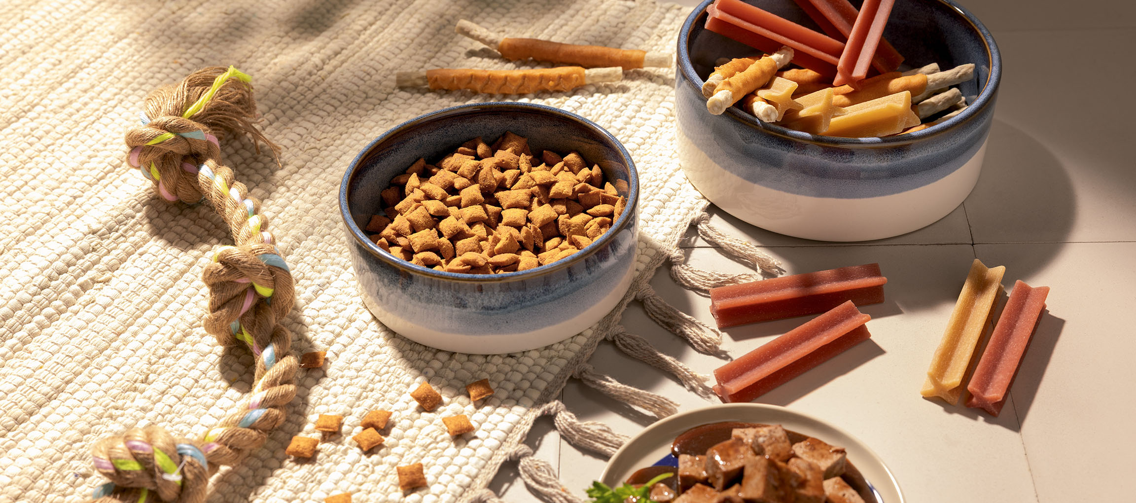 Aliments et friandises variés pour animaux de compagnie, y compris croquettes, bâtonnets à mâcher et jouet en corde.