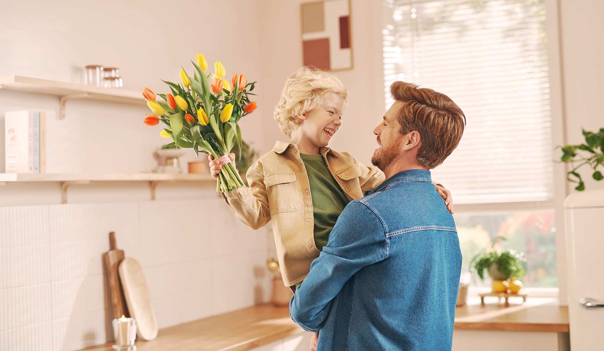 Père tenant son fils qui présente un bouquet de tulipes dans une cuisine lumineuse.