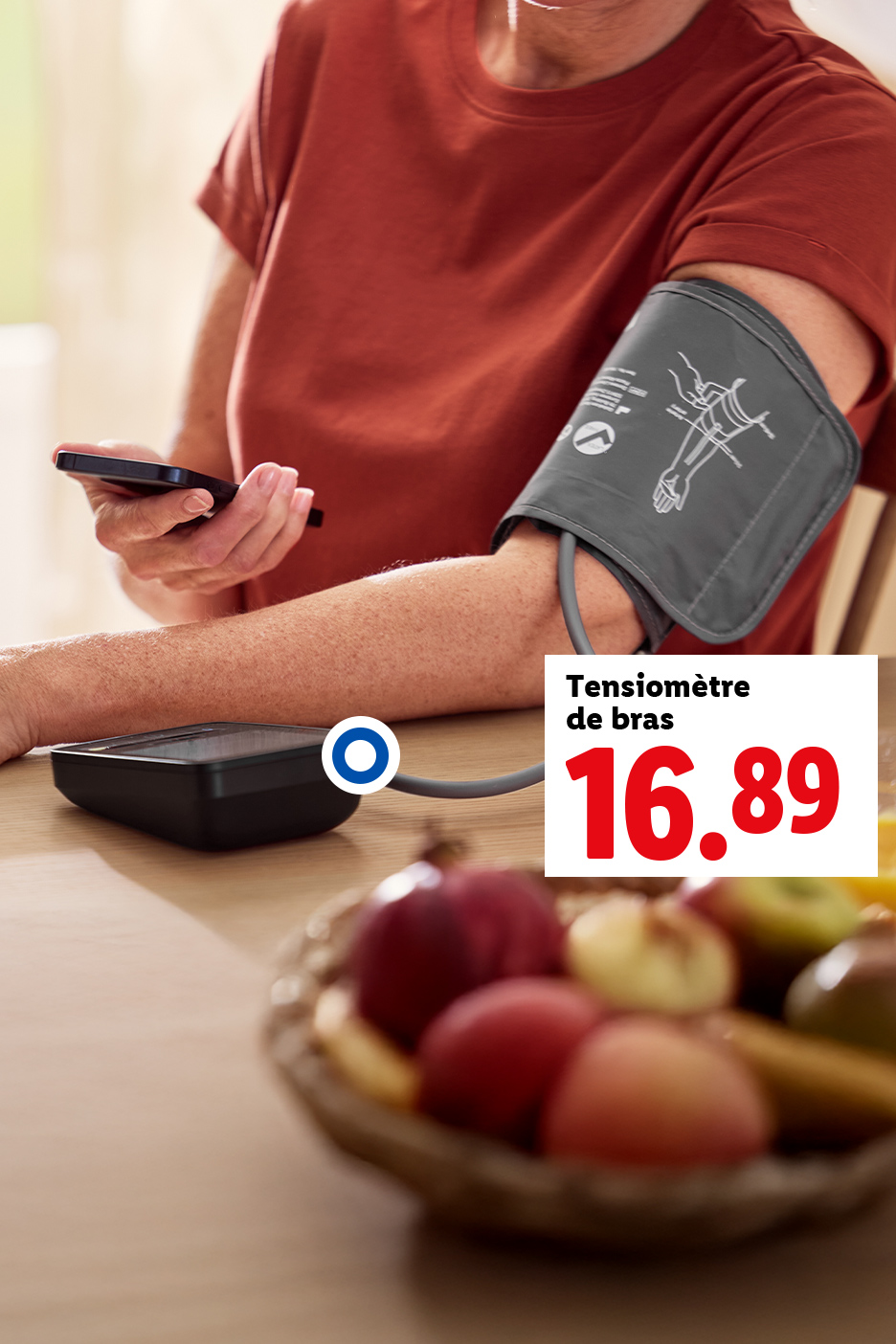 Une personne utilise un tensiomètre de bras et un smartphone, avec un bol de fruits sur la table.