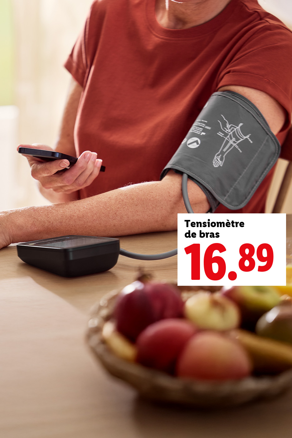 Une personne utilise un tensiomètre de bras et un smartphone, avec un bol de fruits sur la table.