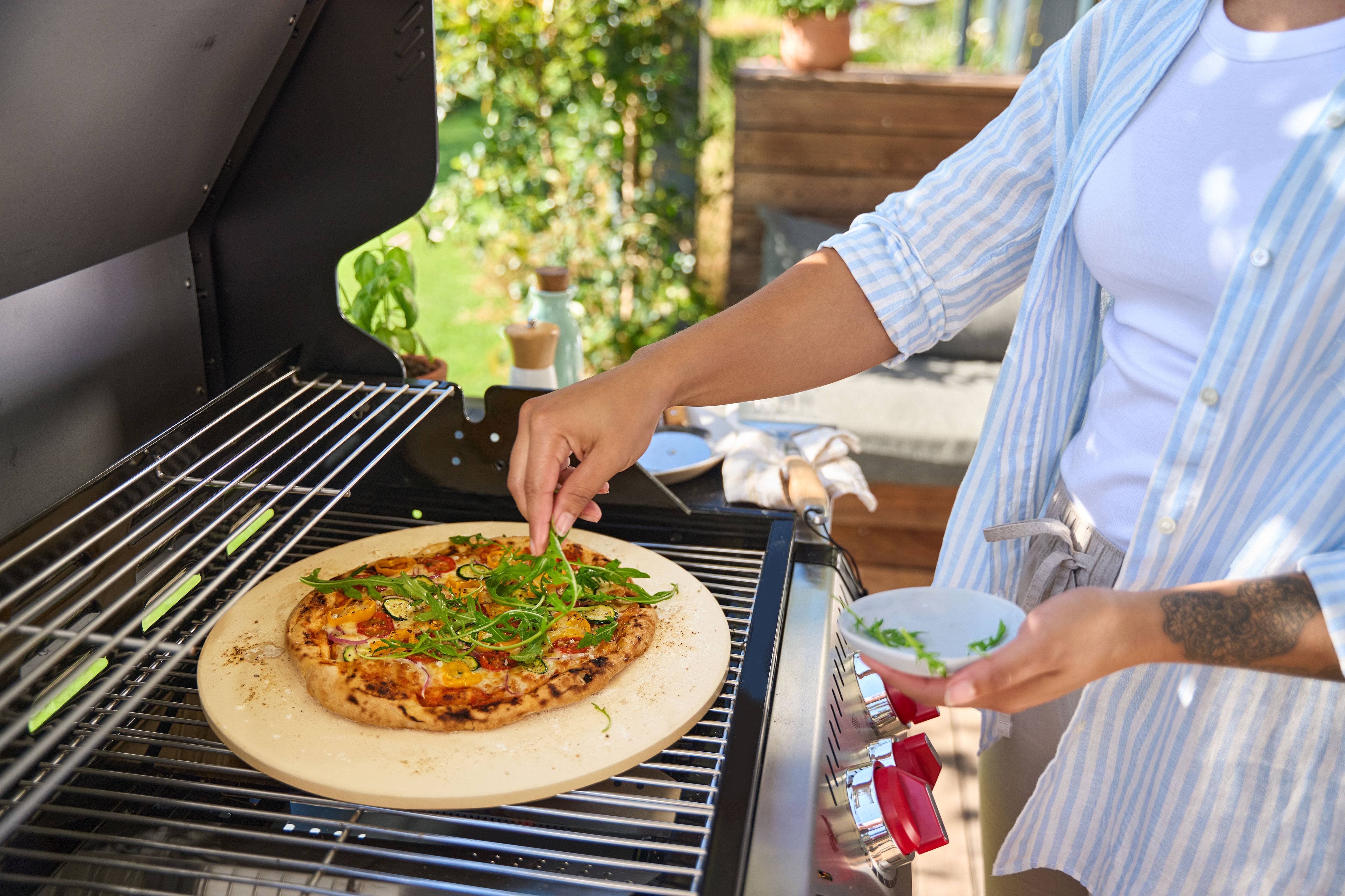 Personne ajoutant de la roquette sur une pizza sur une pierre à pizza dans un barbecue à gaz.