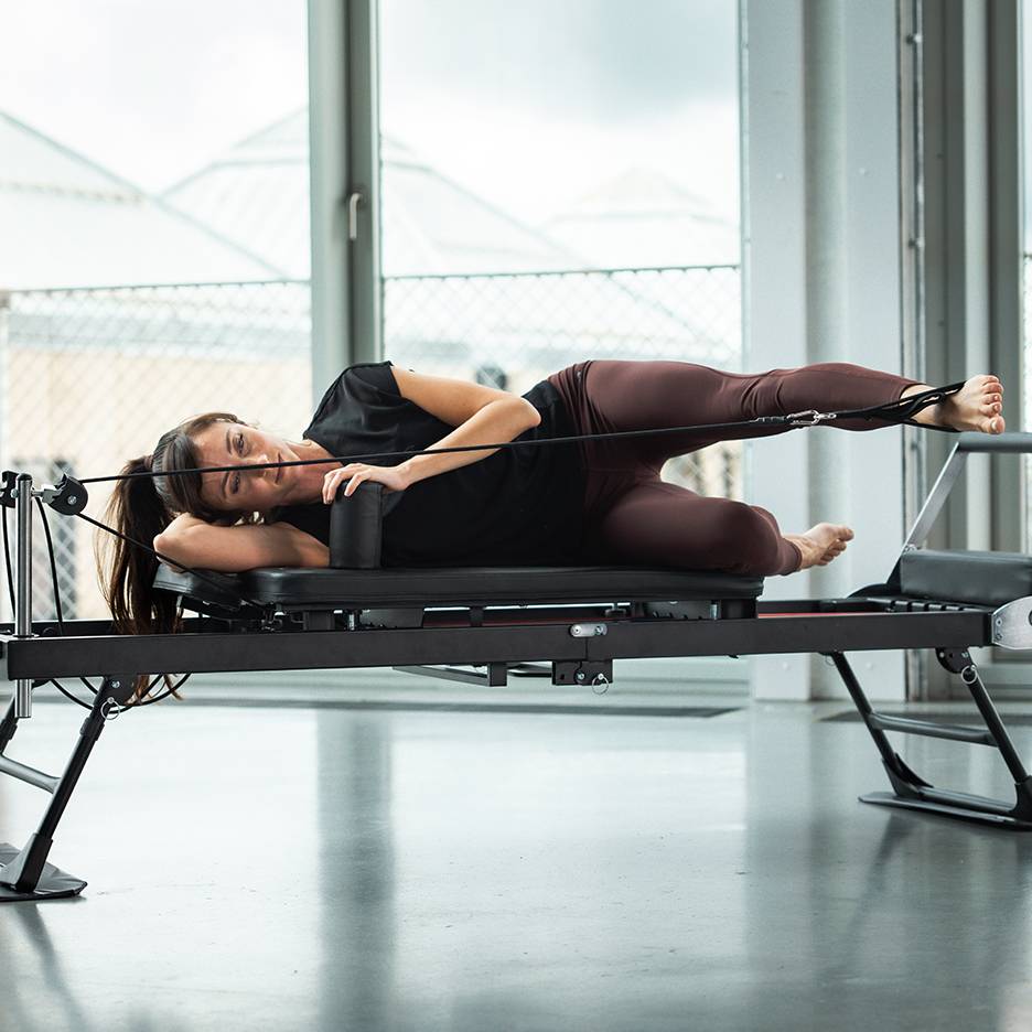 Femme faisant du Pilates sur un reformer, allongée sur le côté avec des sangles aux jambes.