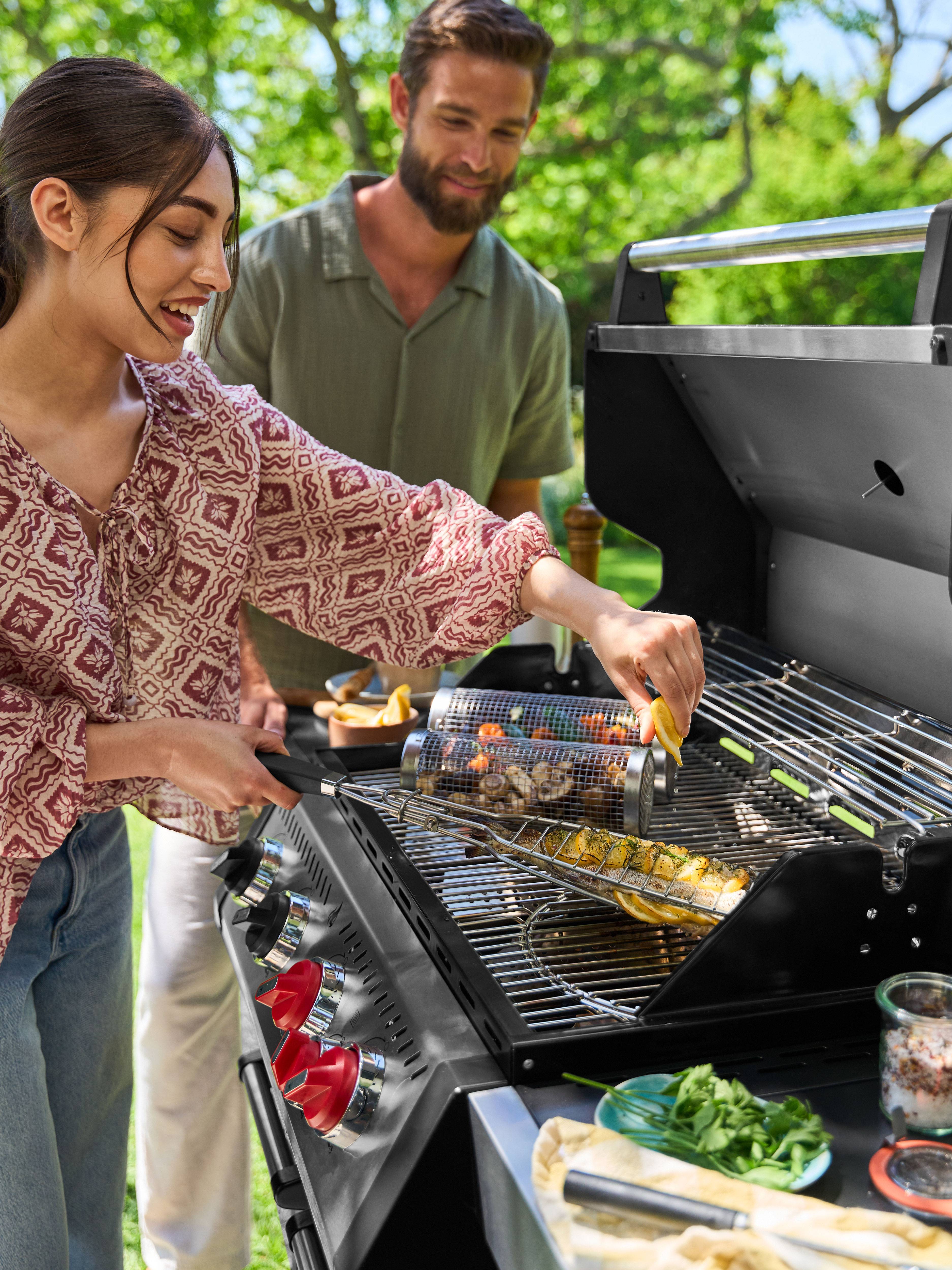 Femme souriante grillant poisson et légumes sur barbecue à gaz, homme regardant de derrière.