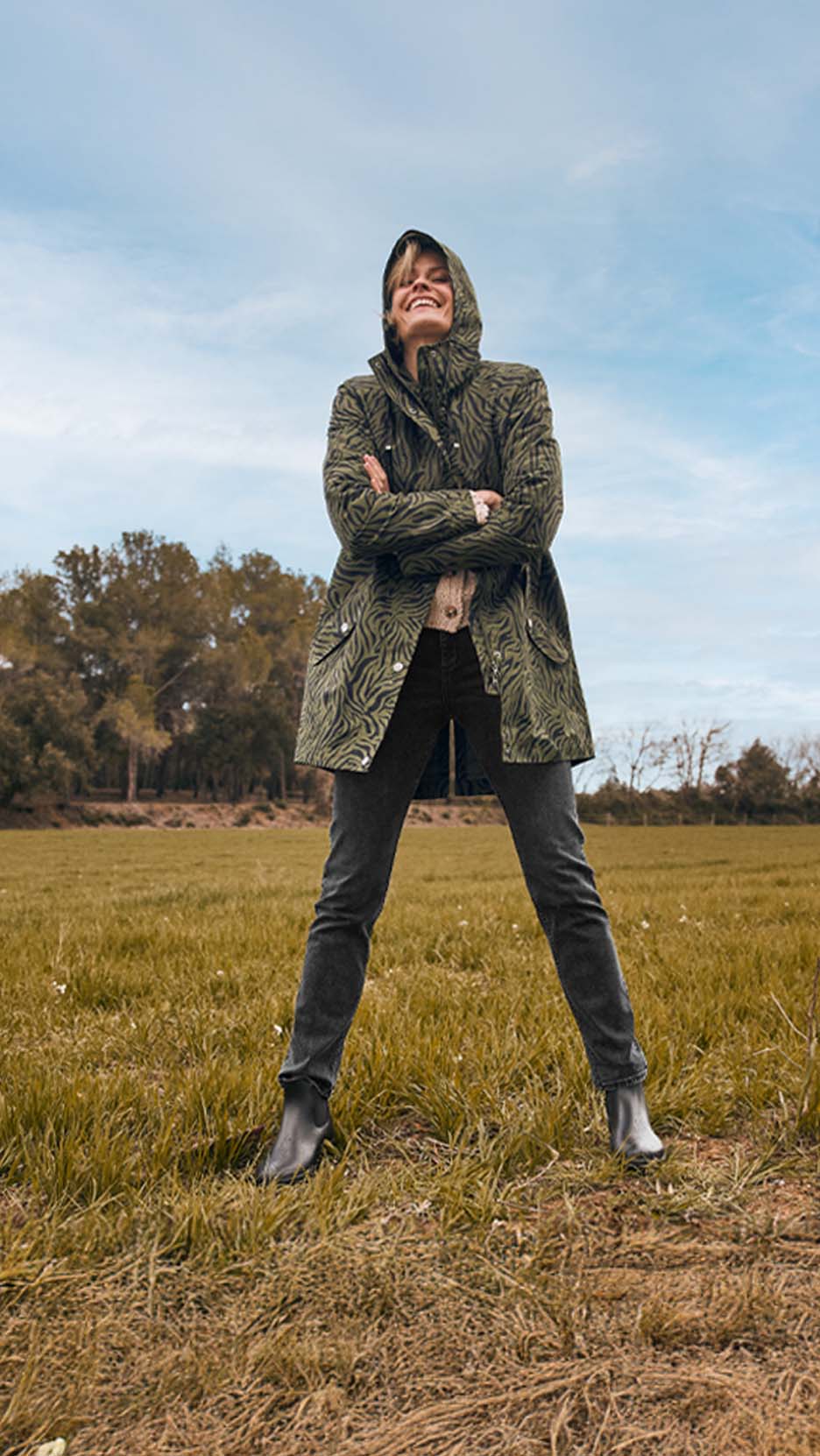 Femme souriante en parka à motif zèbre, jean et bottines dans un champ.