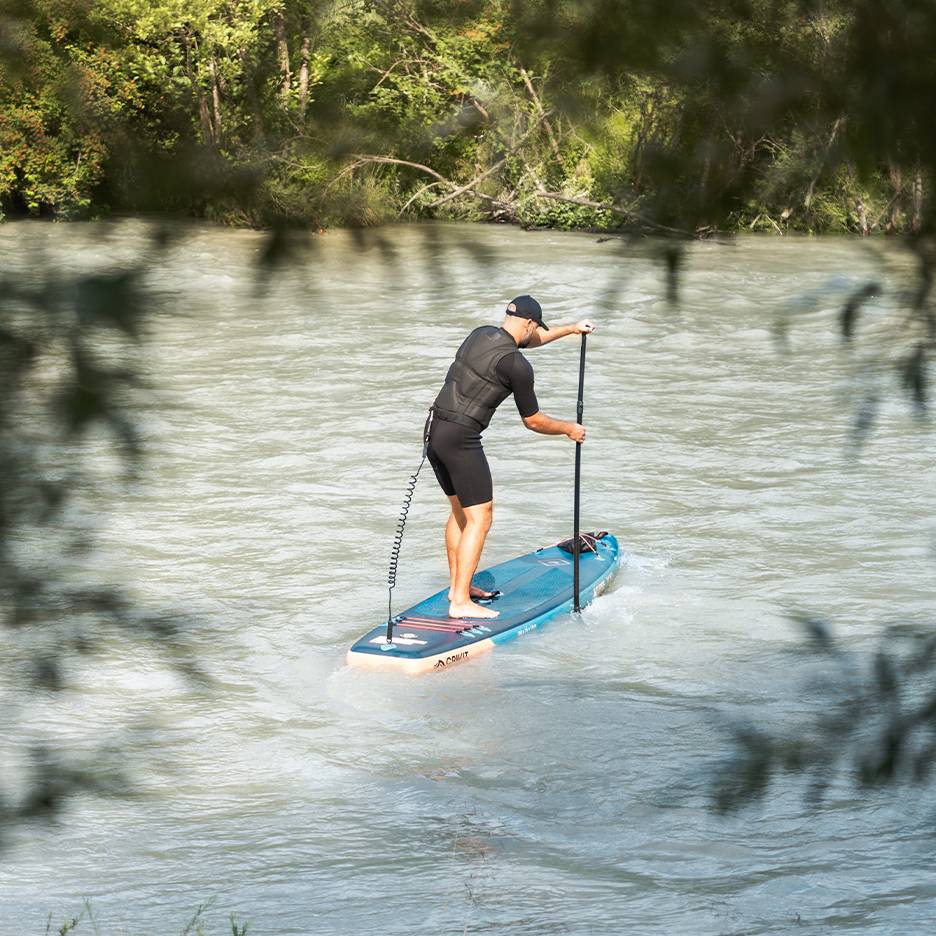 Homme en combinaison et gilet de sauvetage pagayant sur une planche de paddleboard bleue sur une rivière.
