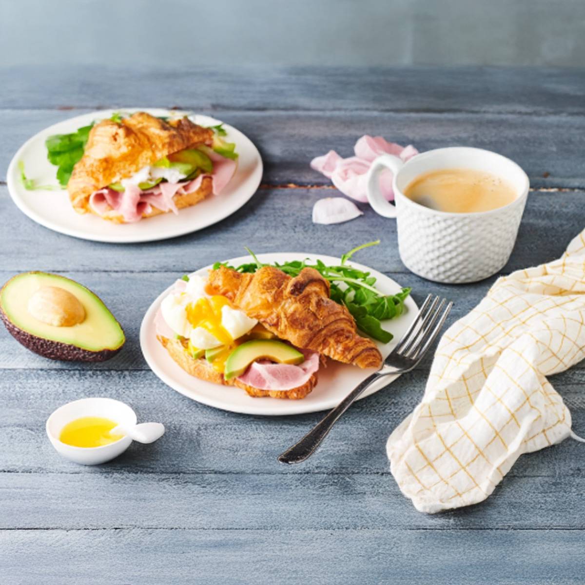 Deux croissants au jambon, avocat et œuf poché, servis avec du café.