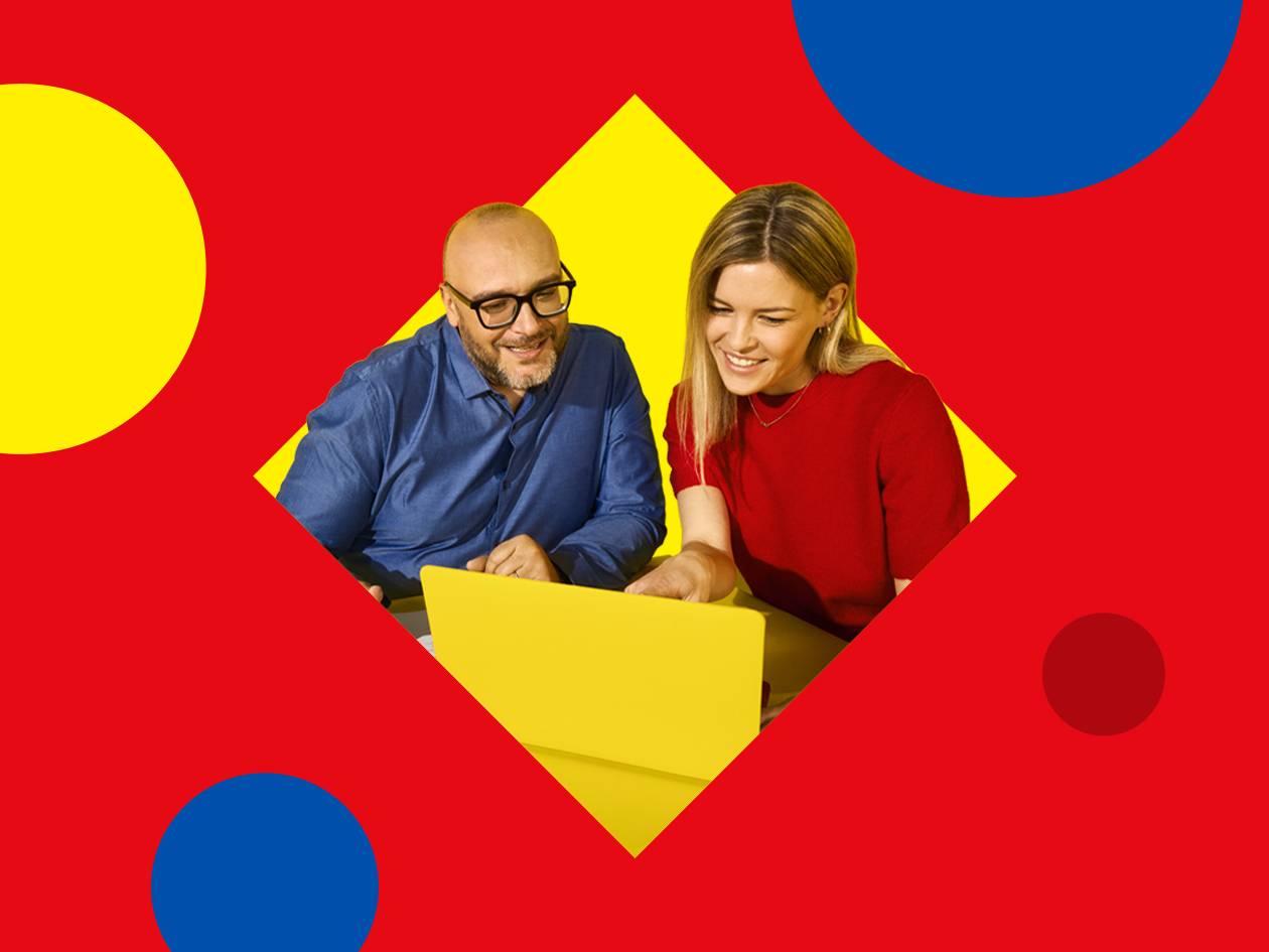 Deux personnes souriantes regardant un ordinateur portable jaune sur un fond rouge avec des formes géométriques.