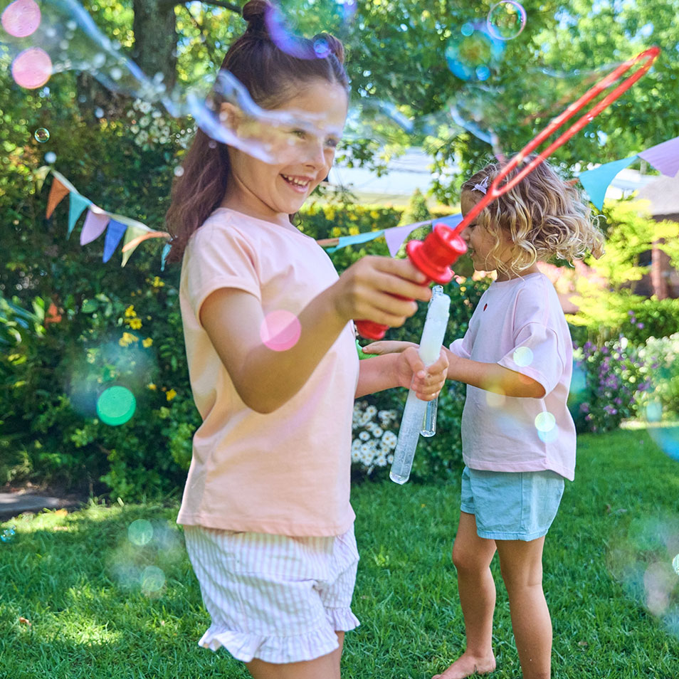 Deux filles jouent avec des baguettes à bulles dans un jardin avec des guirlandes colorées.