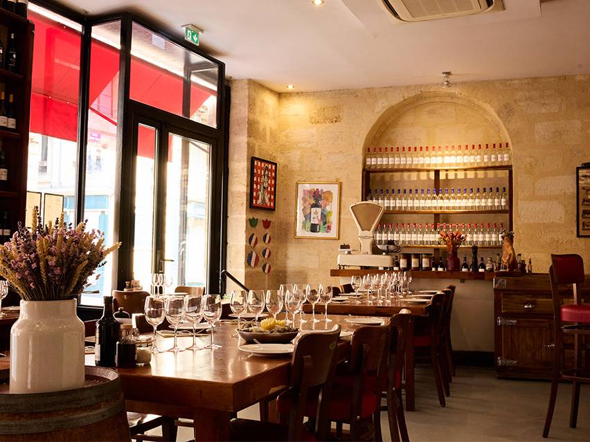 Intérieur de restaurant avec tables en bois, bouteilles de vin sur étagères et grande fenêtre.