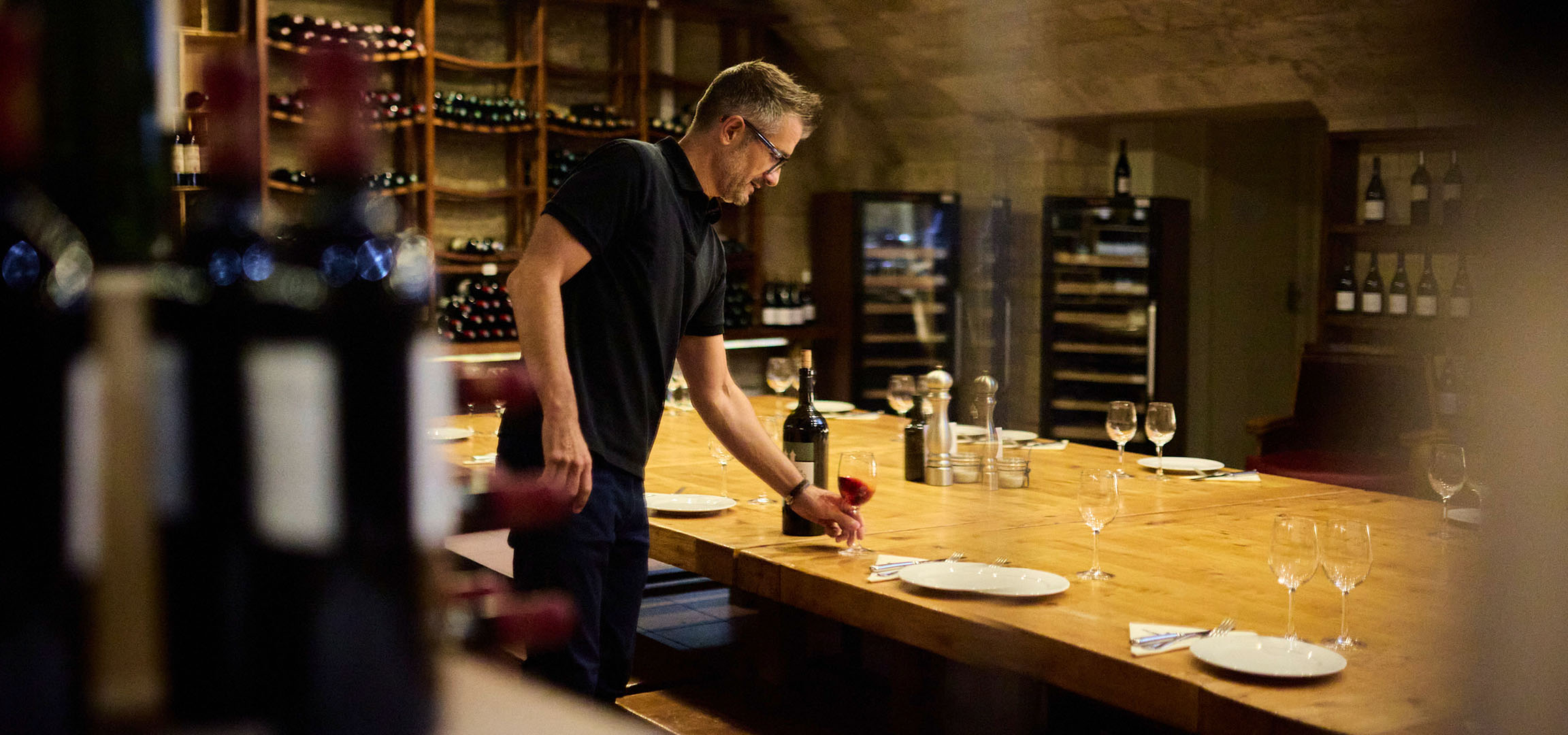 Homme dans une cave à vin, bouteilles de vin sur des étagères et une table.