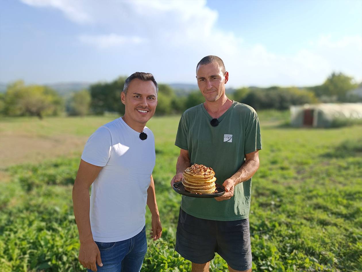 Deux hommes souriants dans un champ, l'un tenant une pile de crêpes.