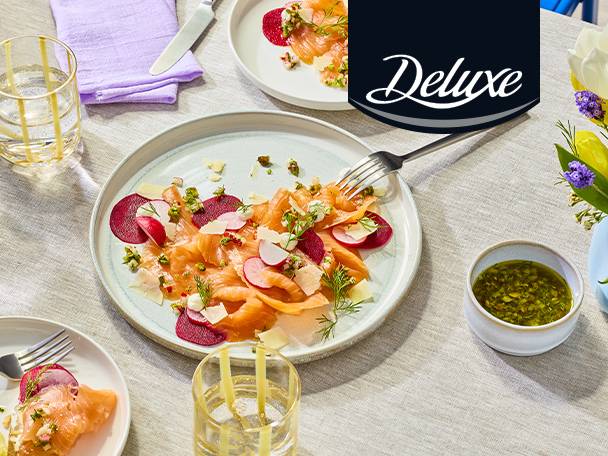 Plat de saumon fumé avec des radis, des betteraves et une sauce verte, avec le logo 'Deluxe'.