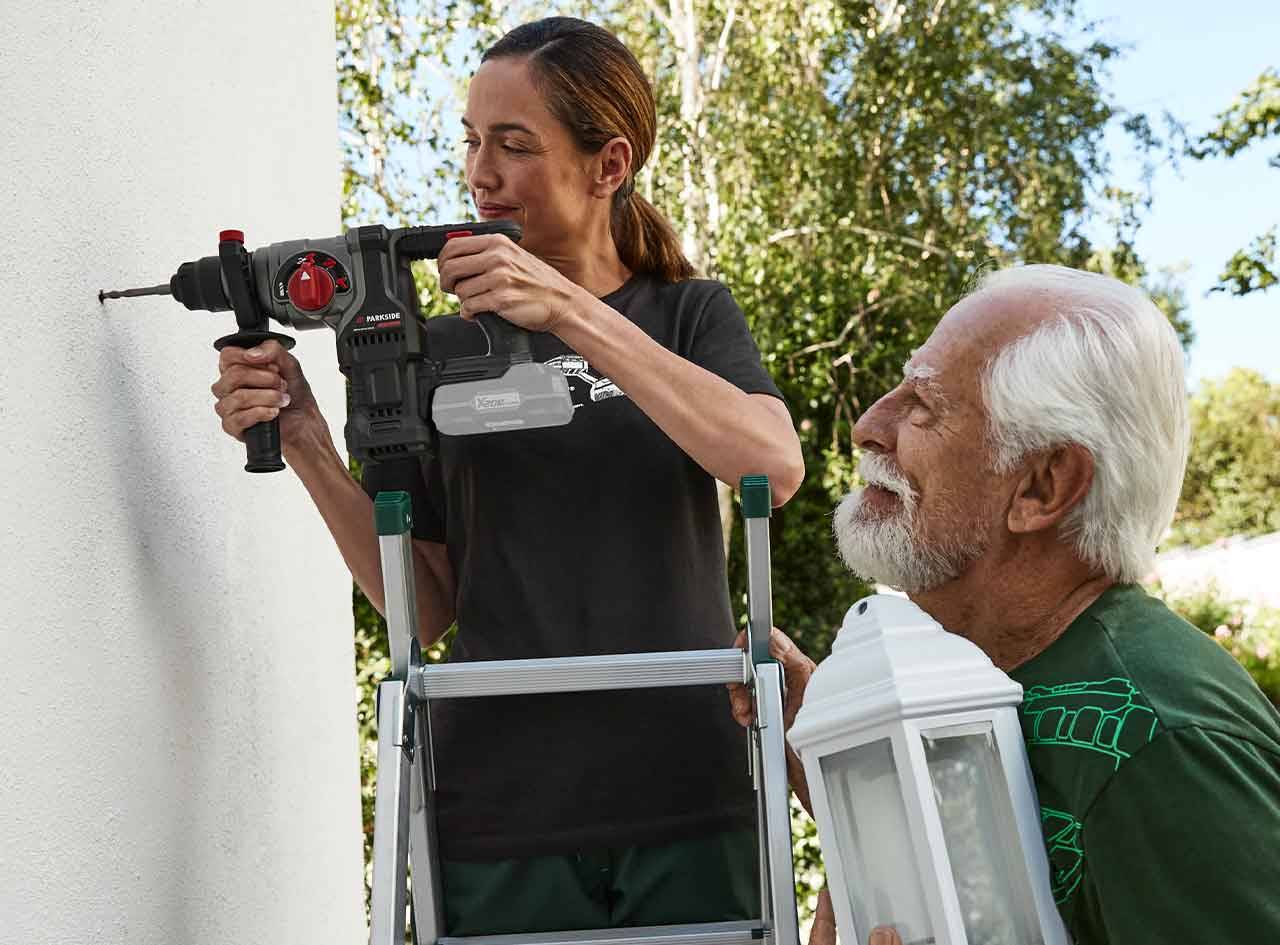 Une femme utilise une perceuse-burineuse Parkside pour fixer un luminaire sur un mur, aidée par un homme.