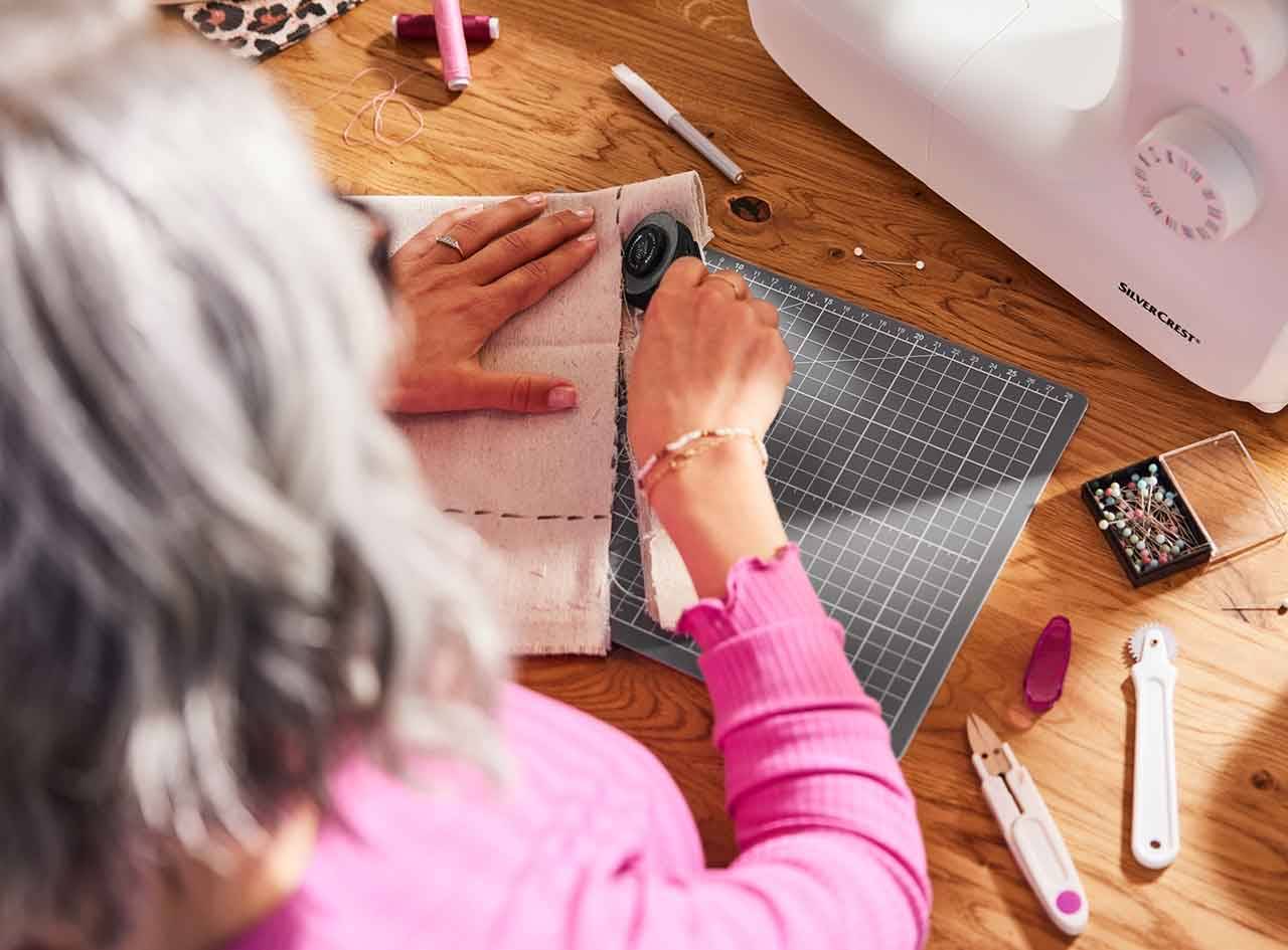Une femme utilise un coupe-tissu rotatif SilverCrest pour couper du tissu sur un tapis de coupe.