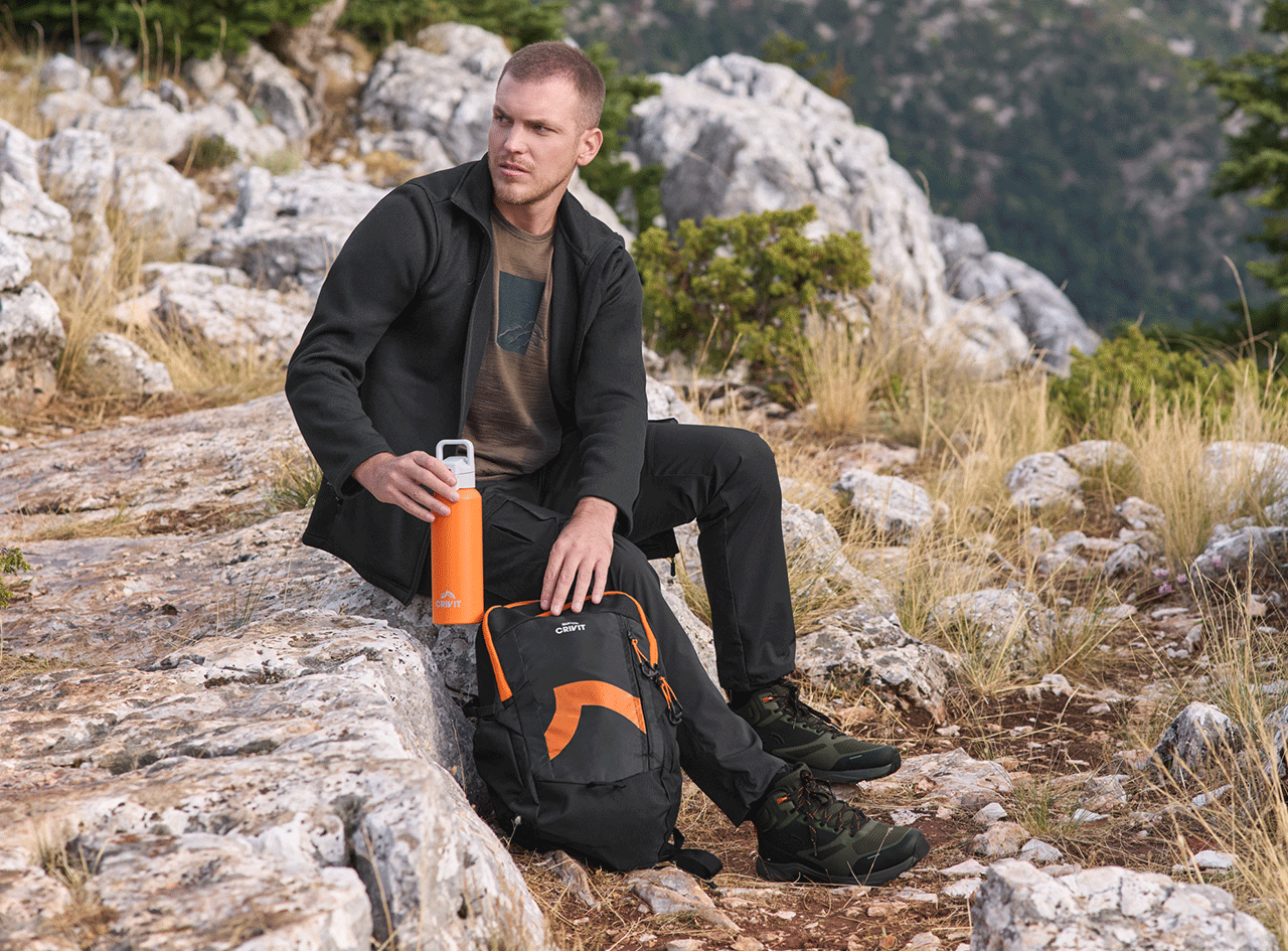 Homme en tenue de randonnée avec gourde orange et sac à dos, assis sur un rocher.
