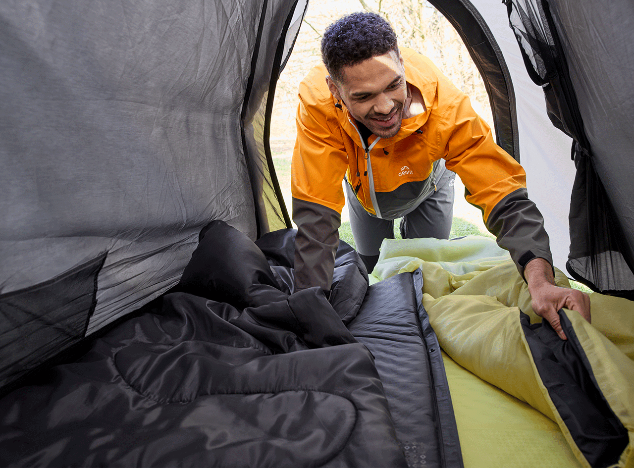 Homme souriant dans une tente, arrangeant un sac de couchage et un matelas de camping.