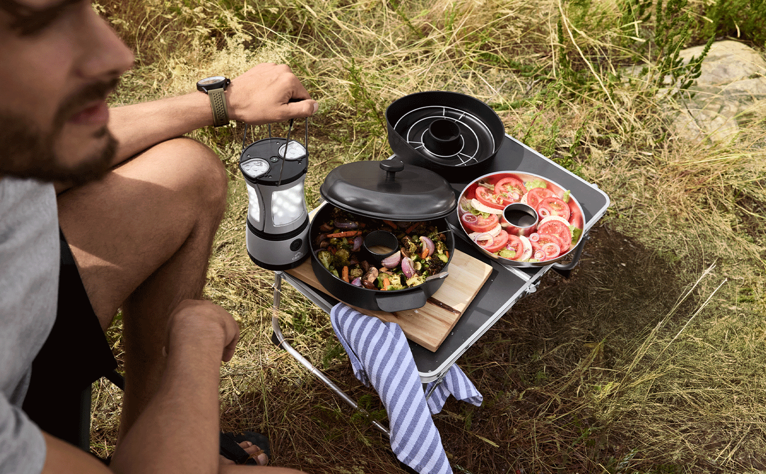 Homme cuisinant en plein air avec une lampe de camping, un plat de légumes rôtis et une salade de tomates.