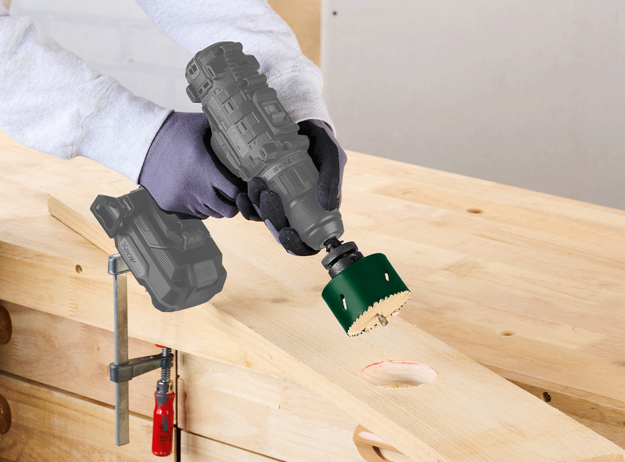 Perceuse sans fil avec scie cloche verte perçant une planche de bois sur un établi.