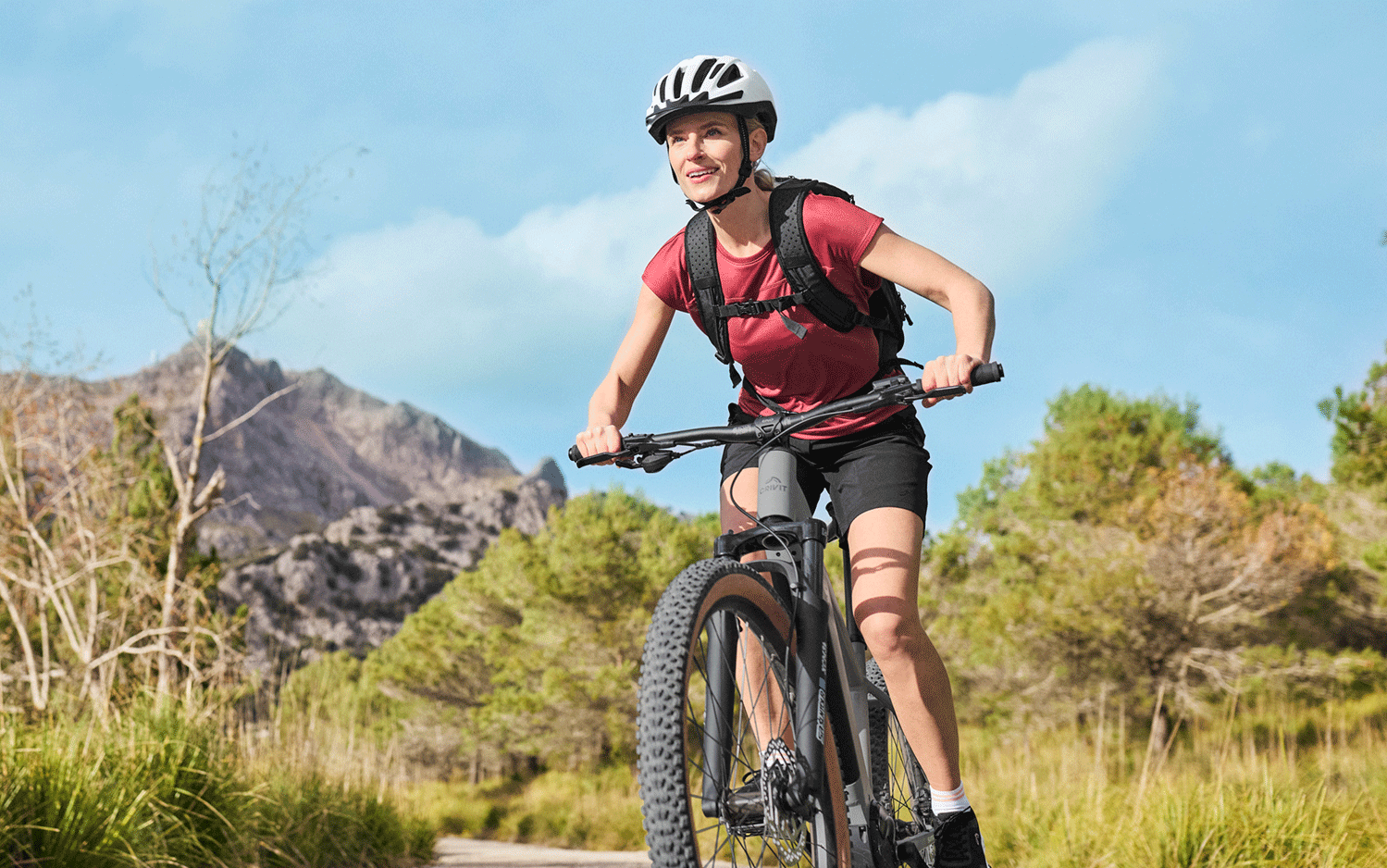 Femme souriante en casque et sac à dos, faisant du VTT sur un sentier de montagne.