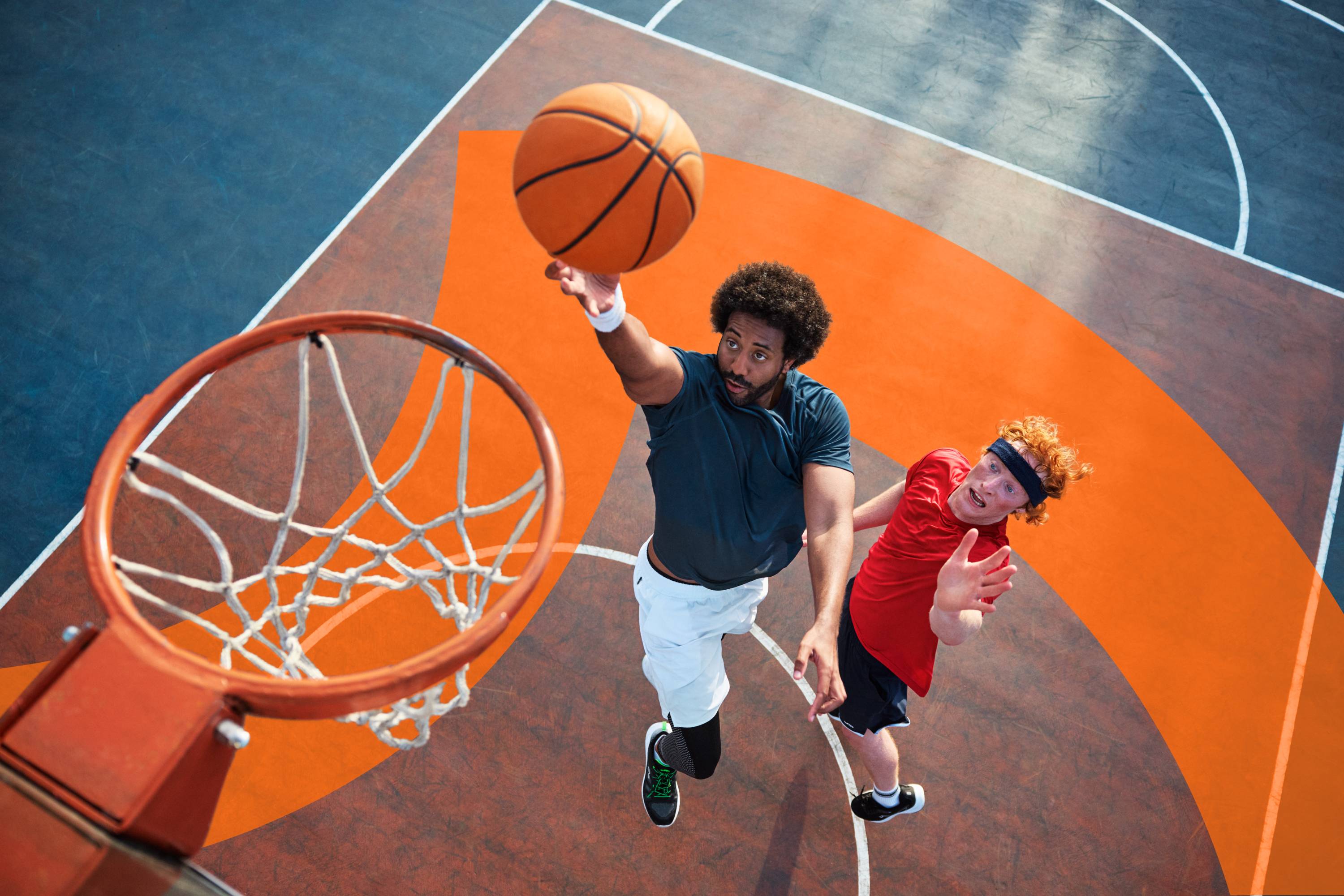 Deux hommes jouent au basketball sur un terrain extérieur, l'un s'apprête à marquer un panier.