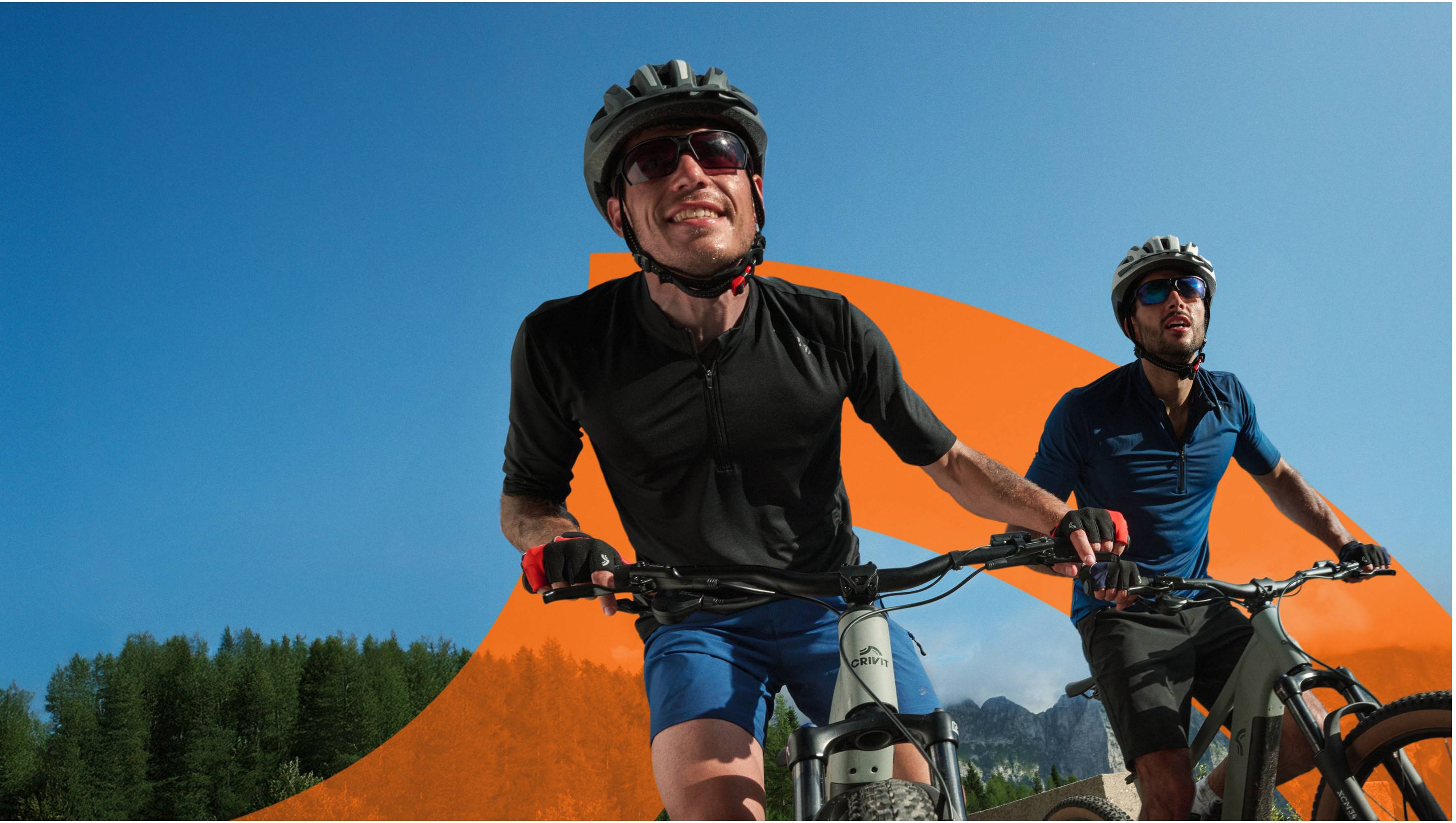 Deux hommes souriants à vélo, portant des casques et des lunettes de soleil, sur un fond de ciel bleu et de montagnes.