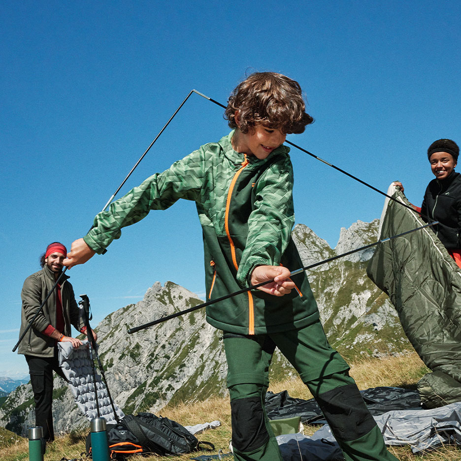 Enfants et adultes montant une tente en montagne, avec des vêtements d'extérieur et des sacs de couchage.