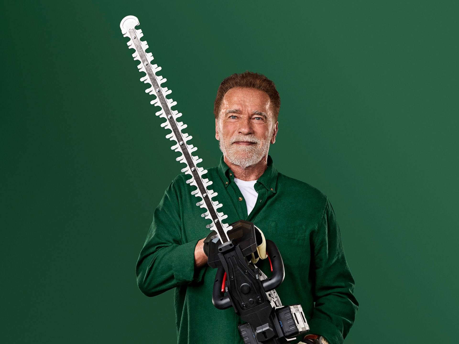 Arnold Schwarzenegger tient un grand taille-haie sur un fond vert.