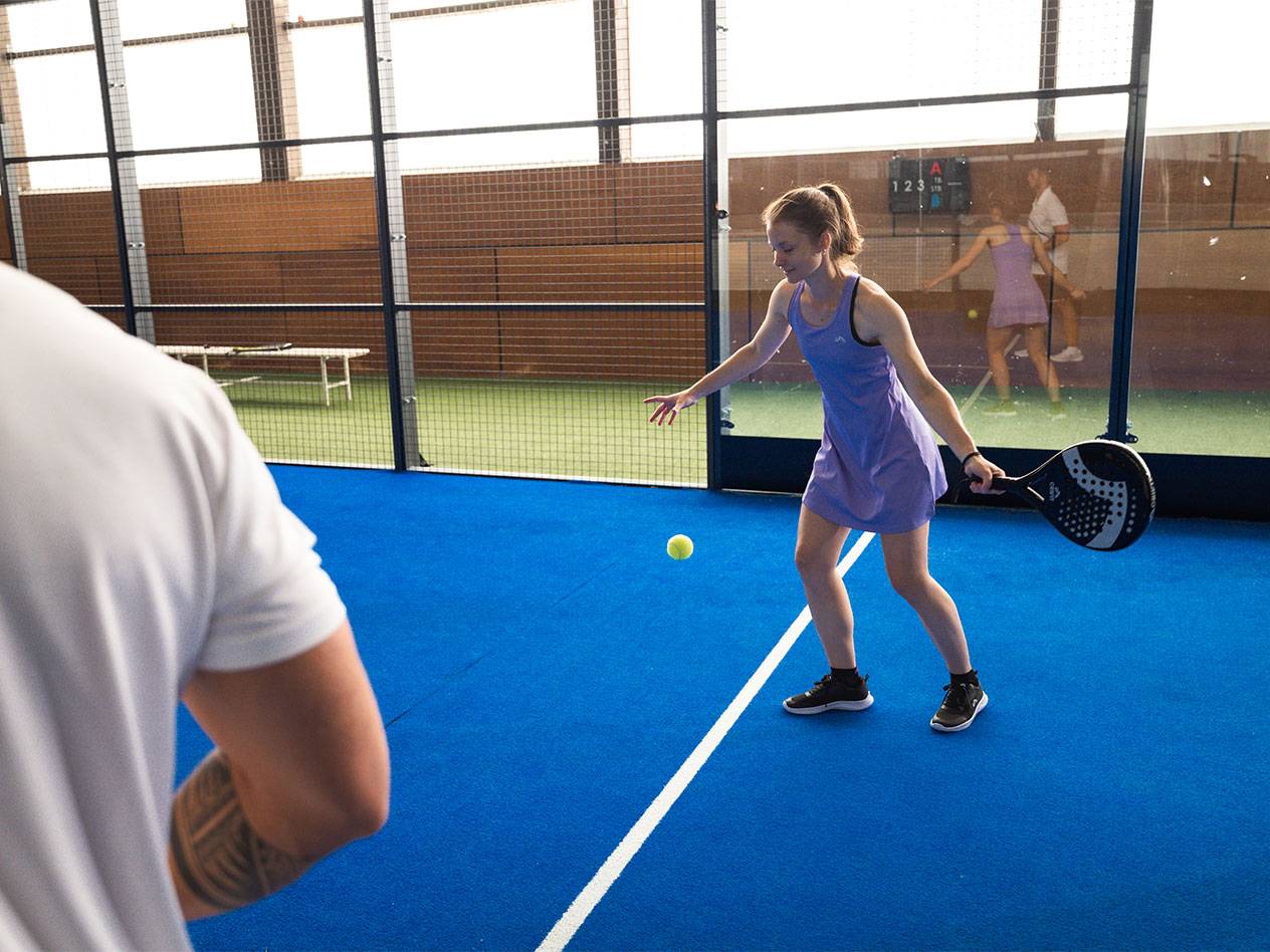 Femme jouant au padel, tenant une raquette et regardant la balle sur un court bleu.