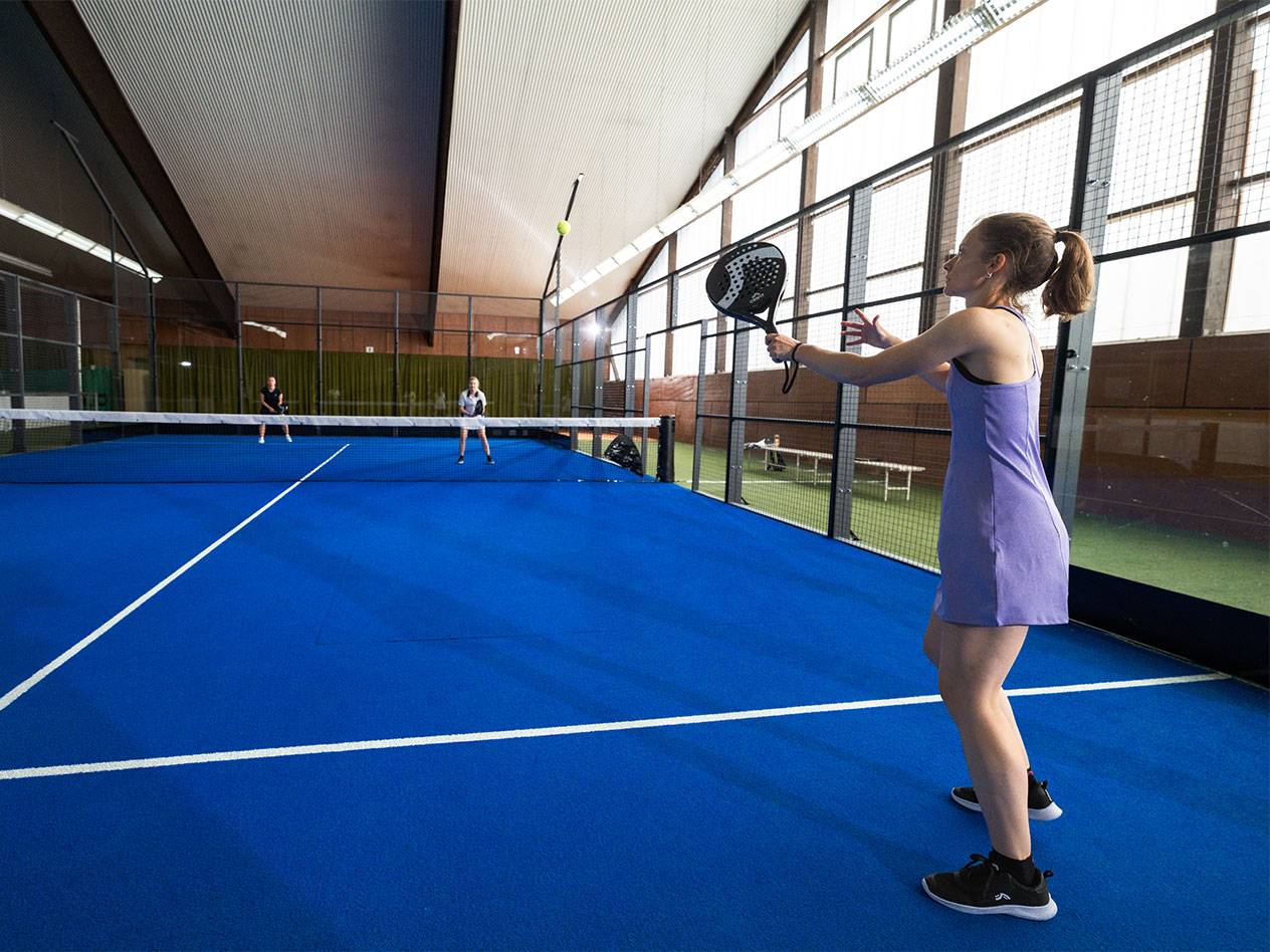 Femme en robe violette jouant au padel sur un court bleu, autres joueurs en arrière-plan.