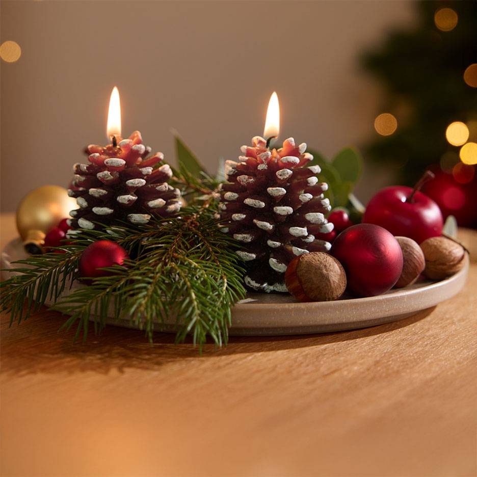 Décoration de l'Avent avec deux bougies en forme de pomme de pin allumées, branches de sapin et décorations de Noël sur un plat.