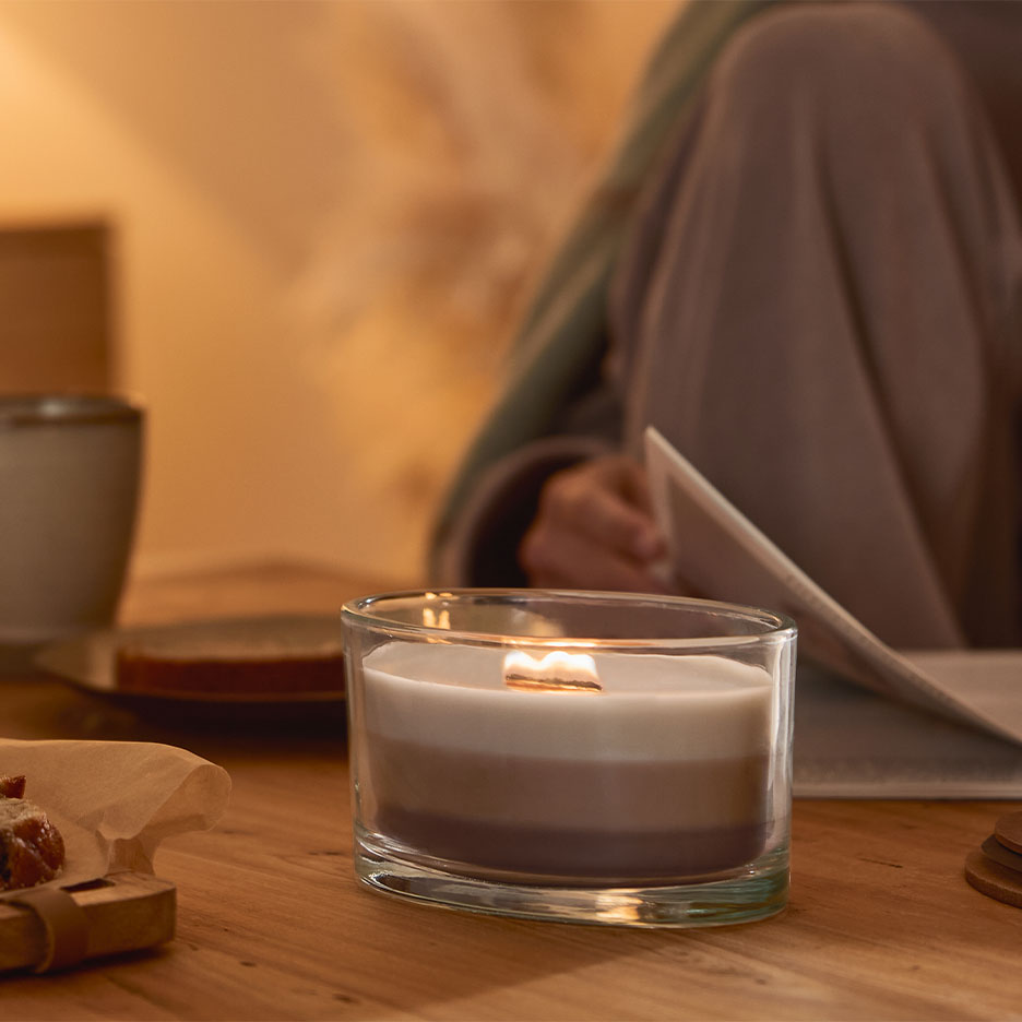 Bougie allumée à trois couches dans un verre, avec une personne lisant en arrière-plan.