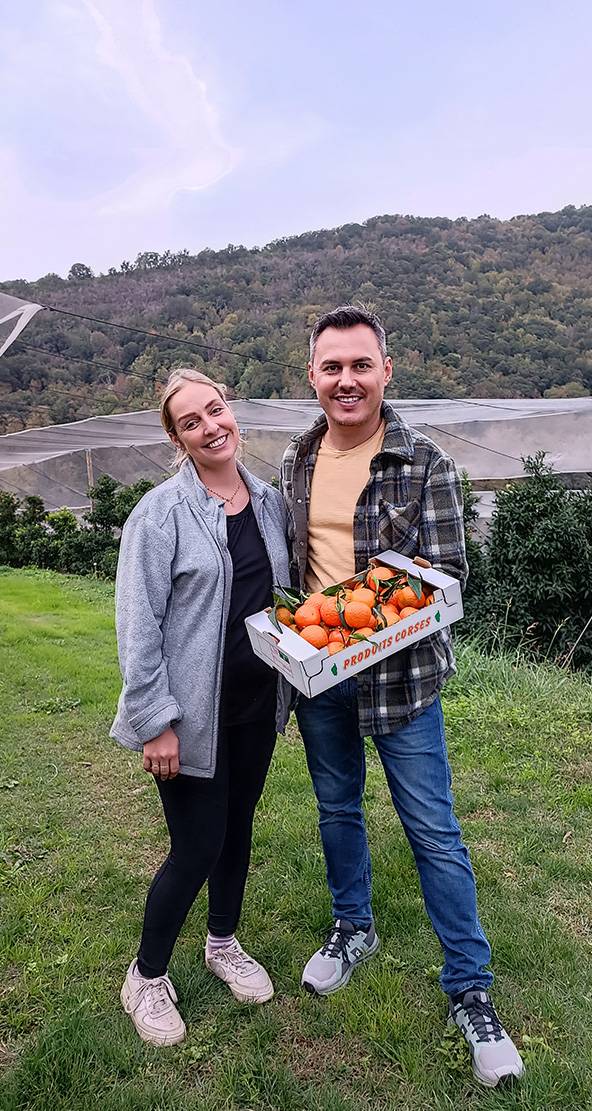 Un homme et une femme souriants tenant une boîte de mandarines corses dans un verger.