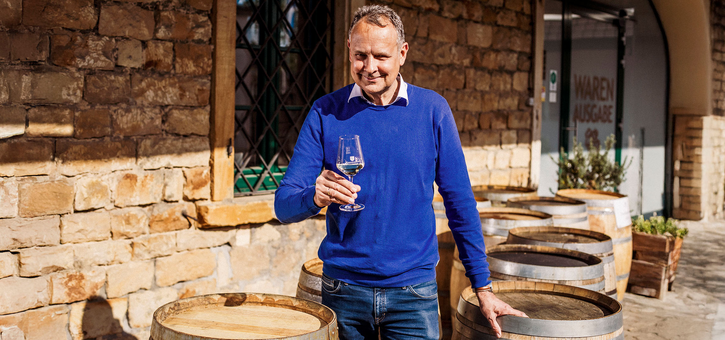 Homme souriant en pull bleu tenant un verre de vin blanc devant un mur de pierre.