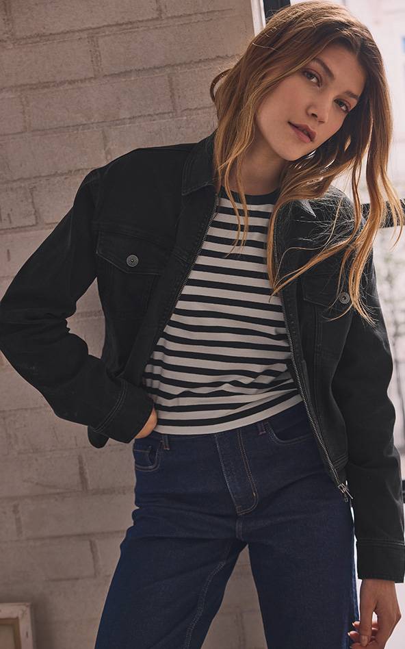 Une femme porte une veste en jean noire, un t-shirt rayé et un jean foncé.