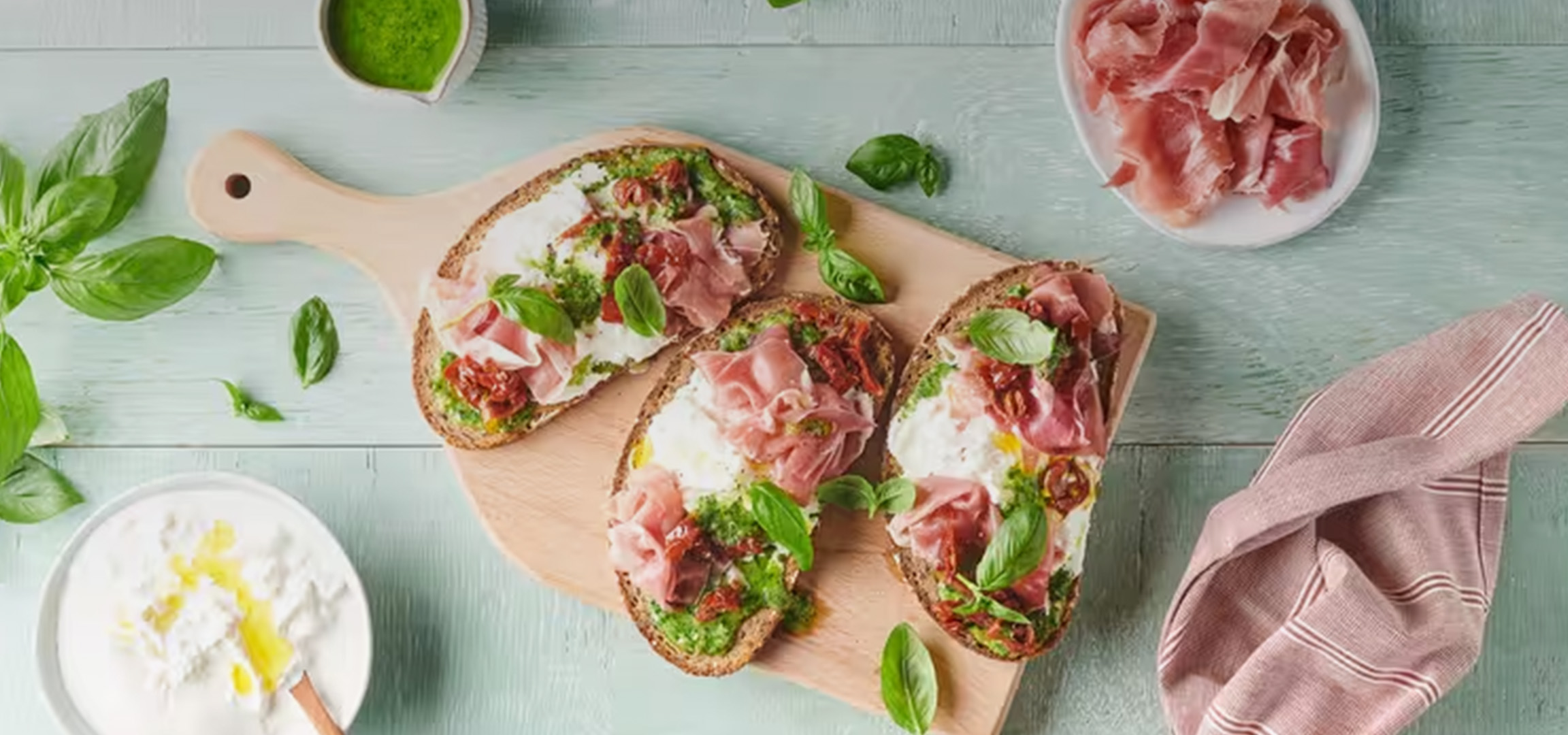 Bruschetta au pesto, jambon cru, tomates séchées et basilic frais sur une planche en bois.