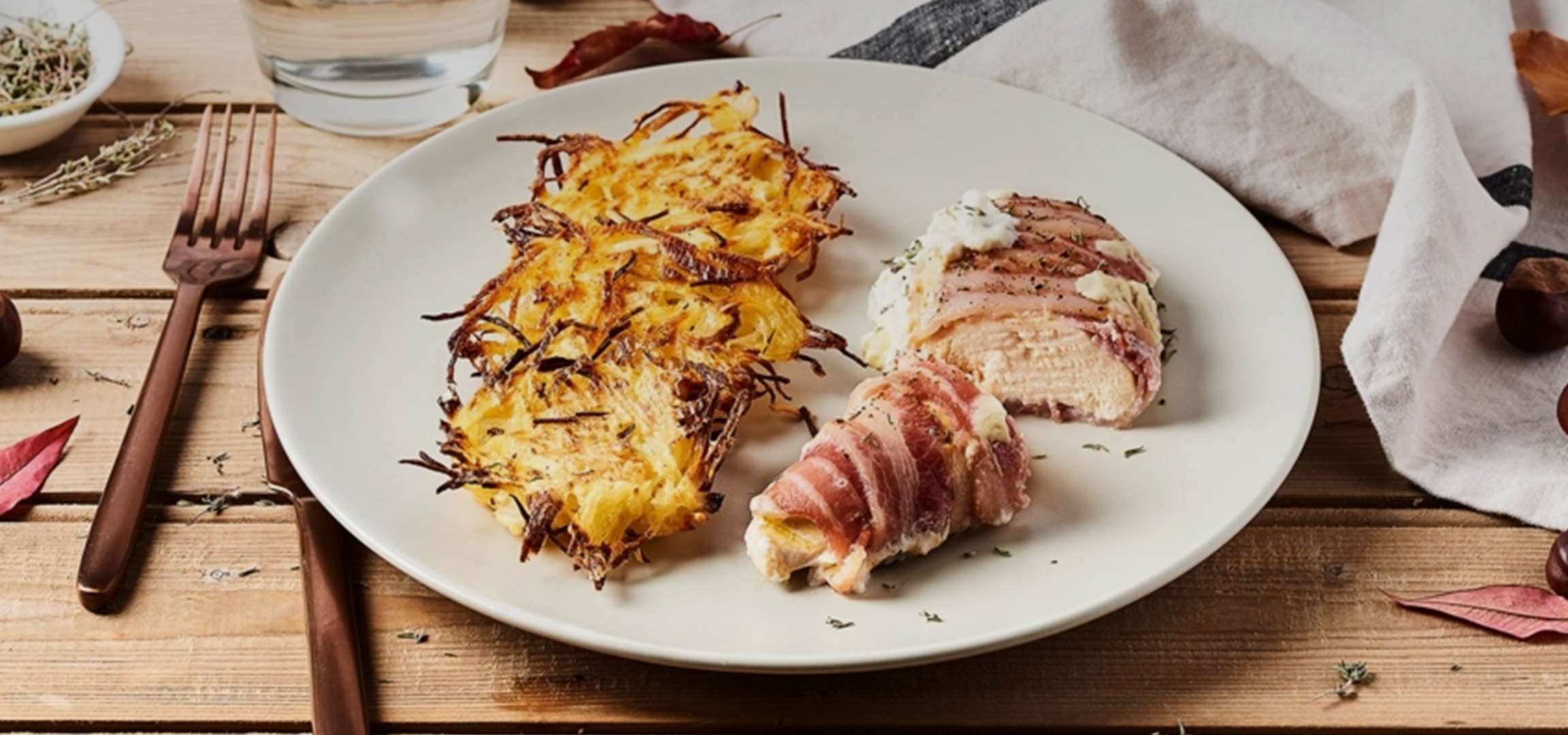 Röstis dorés et poulet enroulé dans du bacon sur une assiette blanche, avec des châtaignes et une serviette.