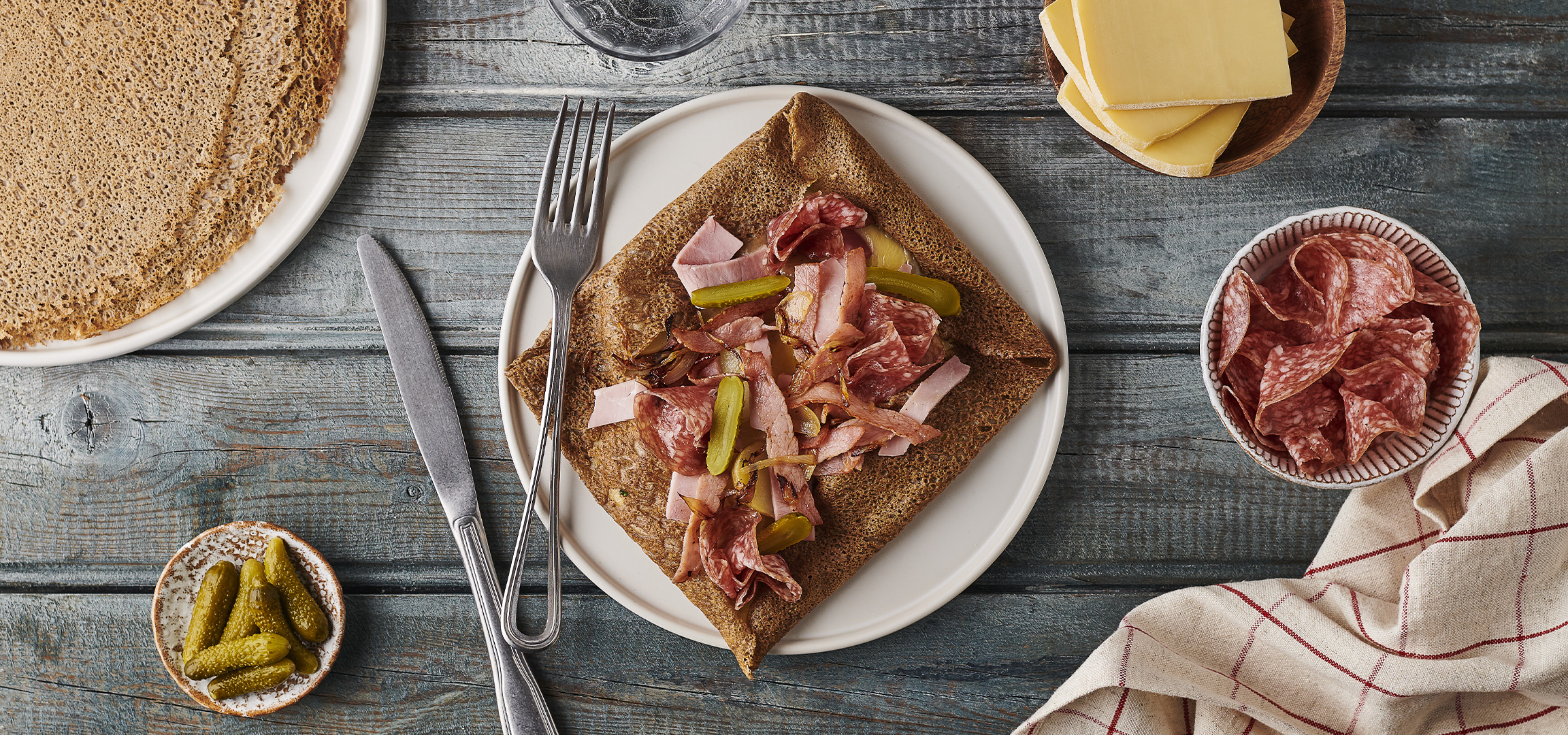 Galette de sarrasin garnie de charcuterie, cornichons et oignons caramélisés, avec un bol de salami.
