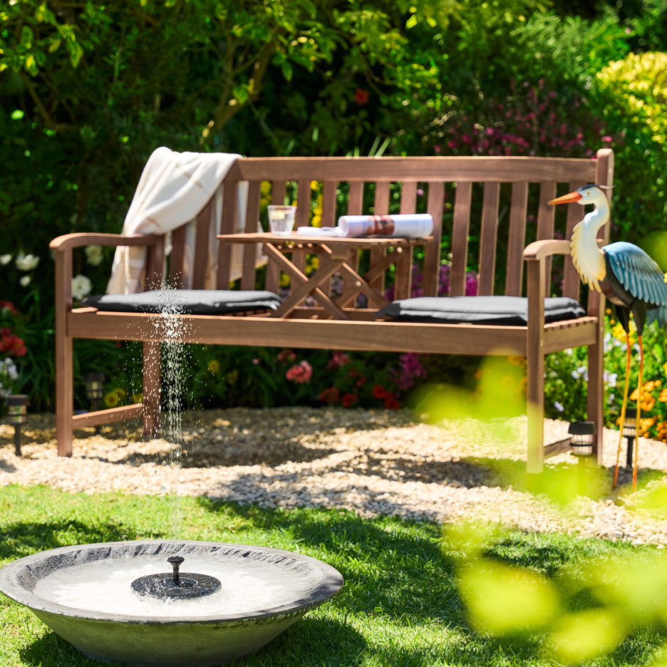 Banc de jardin en bois avec table pliante, coussins et fontaine solaire dans un jardin ensoleillé.