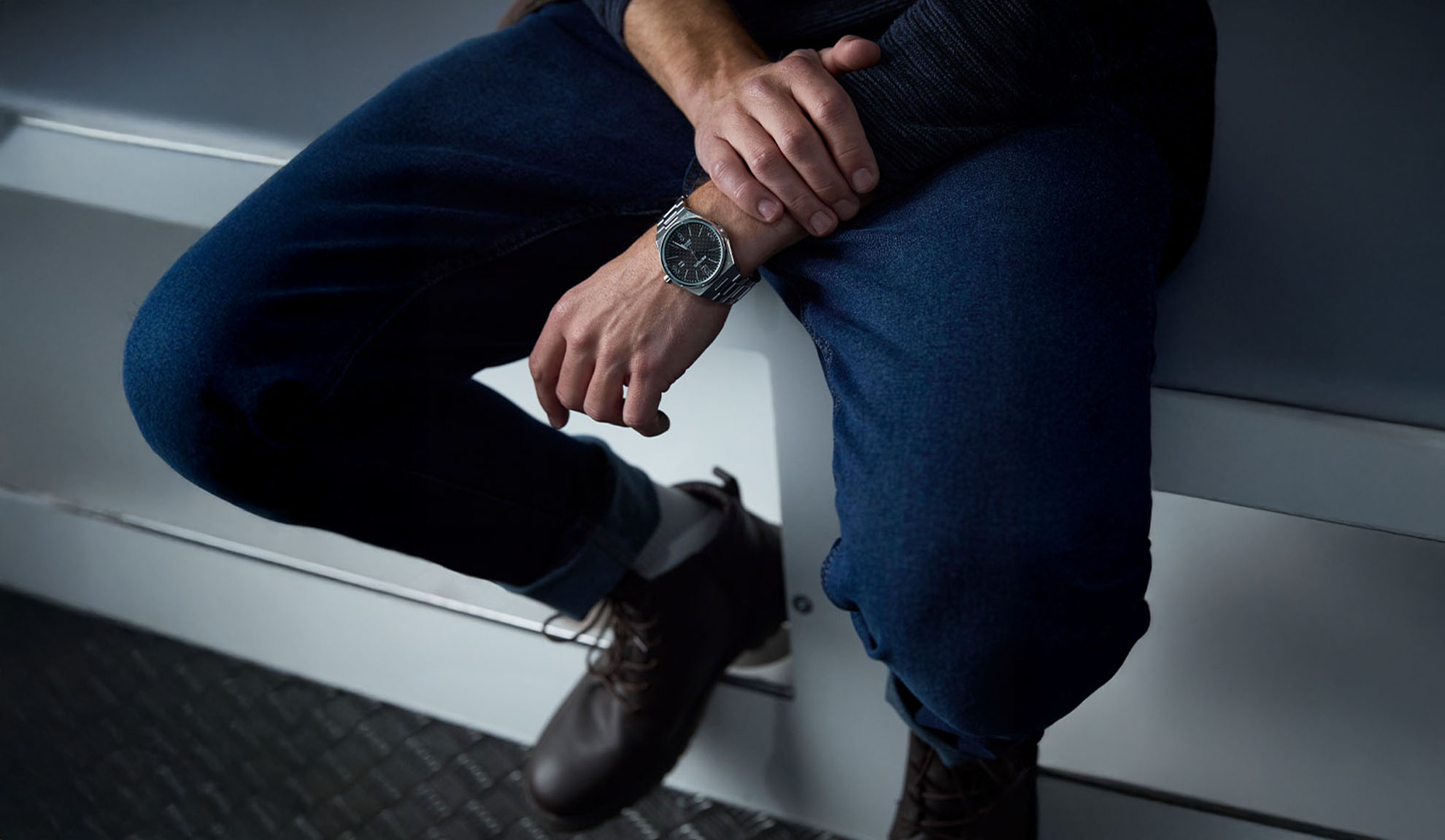 Homme portant un jean bleu foncé, un pull sombre et une montre argentée, assis.