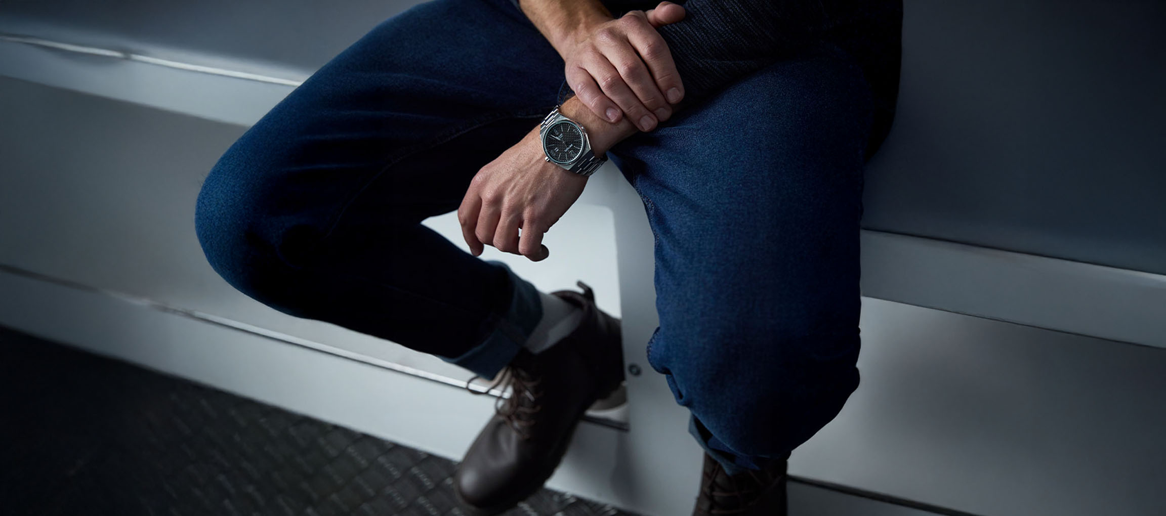 Homme en jean bleu, bottes marron et montre argentée, assis sur une surface blanche.