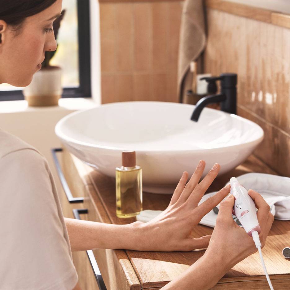 Femme utilisant un set électrique de manucure/pédicure sur une coiffeuse en bois avec un lavabo blanc.