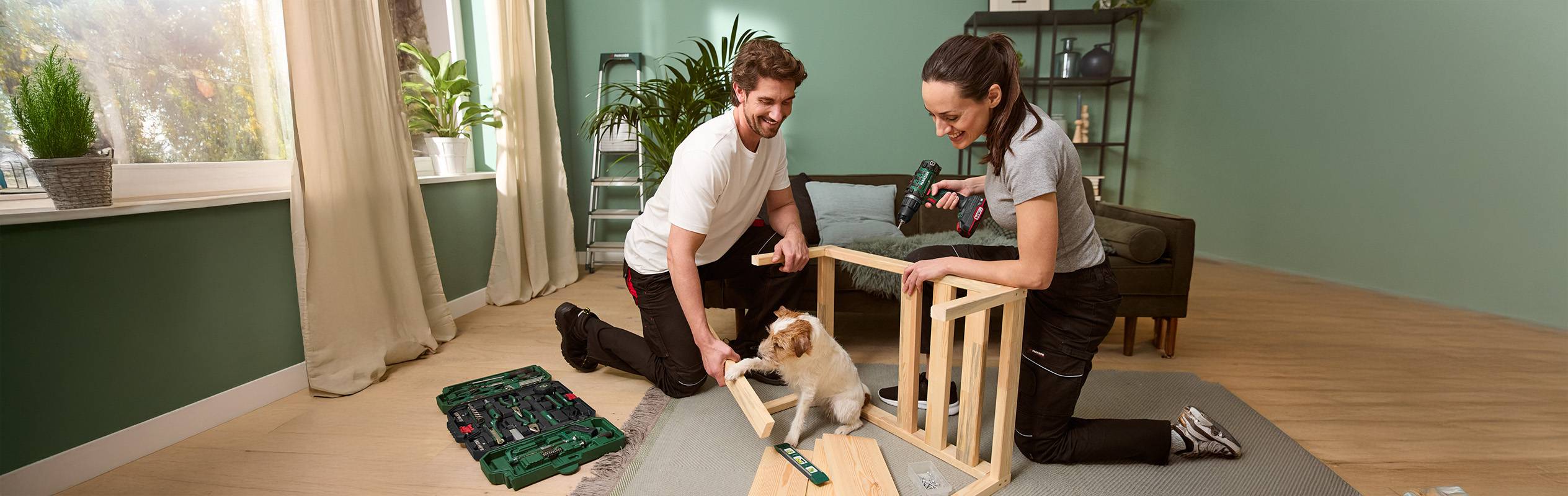Un couple assemble un meuble en bois avec un tournevis Bosch, leur chien les observant.