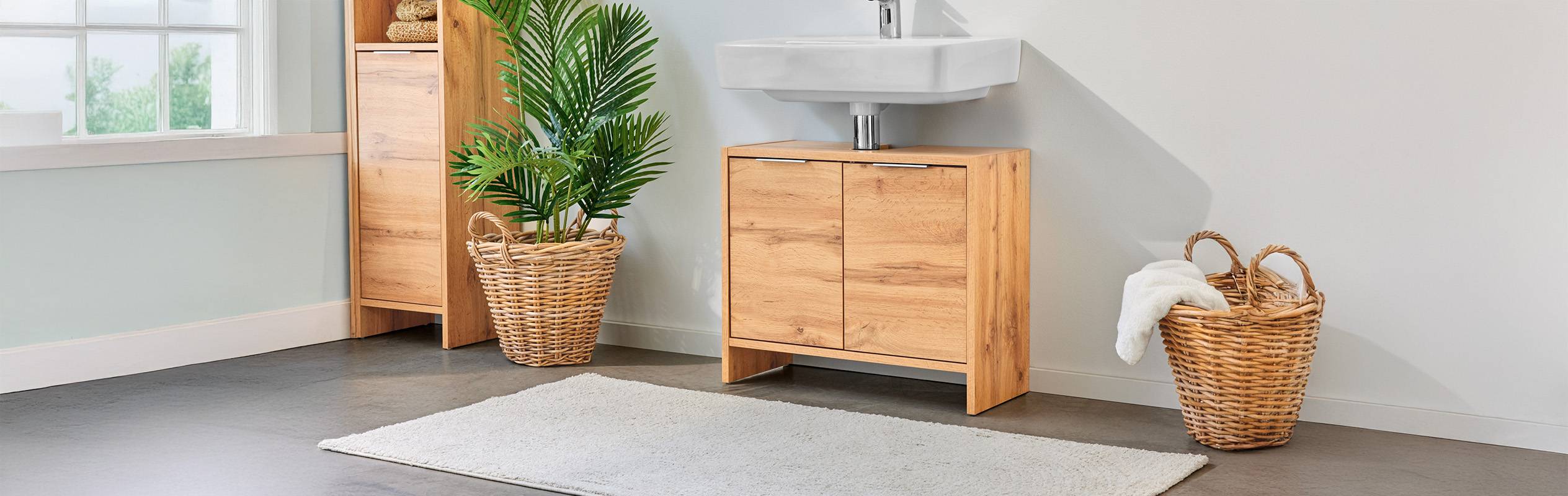 Meuble de salle de bain en bois clair avec lavabo, paniers en osier et tapis.