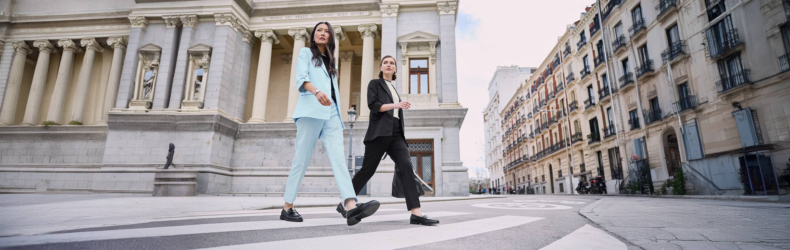 Deux femmes en costumes élégants traversant une rue de ville devant un bâtiment classique.