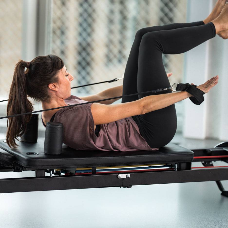 Femme faisant du Pilates sur un reformer, vêtue de vêtements de sport noirs.
