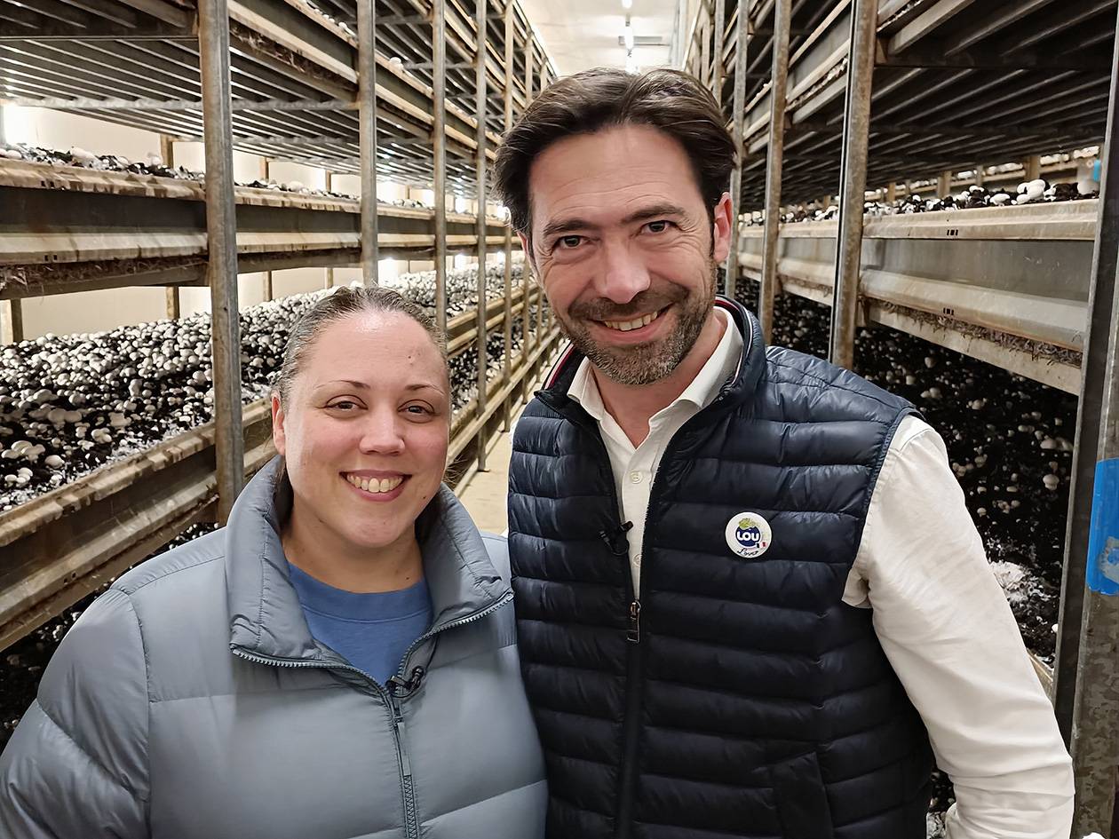 Deux personnes souriantes dans une champignonnière, avec des champignons blancs sur des étagères.